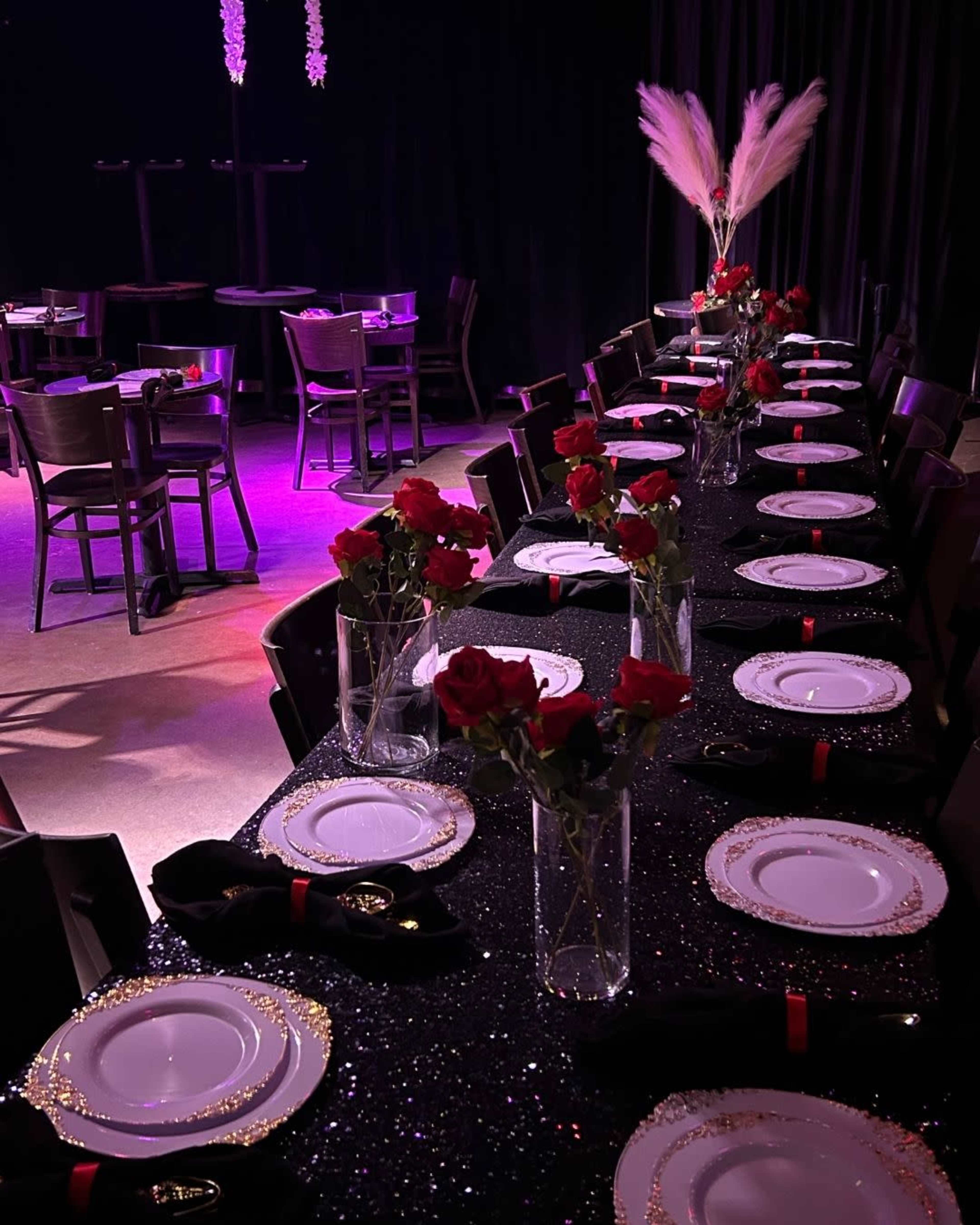 A long dining table is elegantly set with white plates, black napkins, and vases of red roses, beneath a dimly lit atmosphere adorned with decorative lighting.
