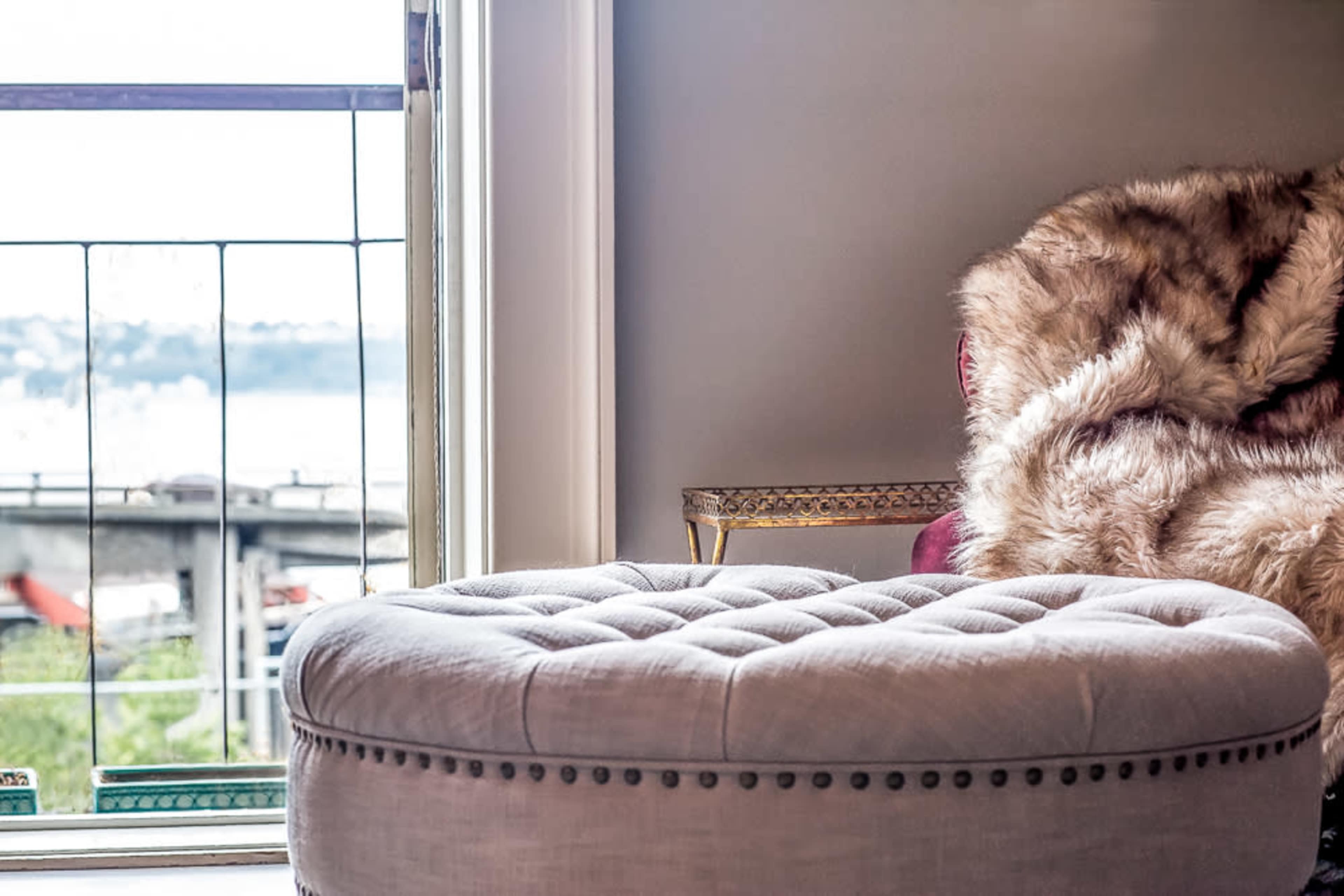 A tufted ottoman is positioned near a window, with a plush chair draped in fur nearby, overlooking a waterfront view.