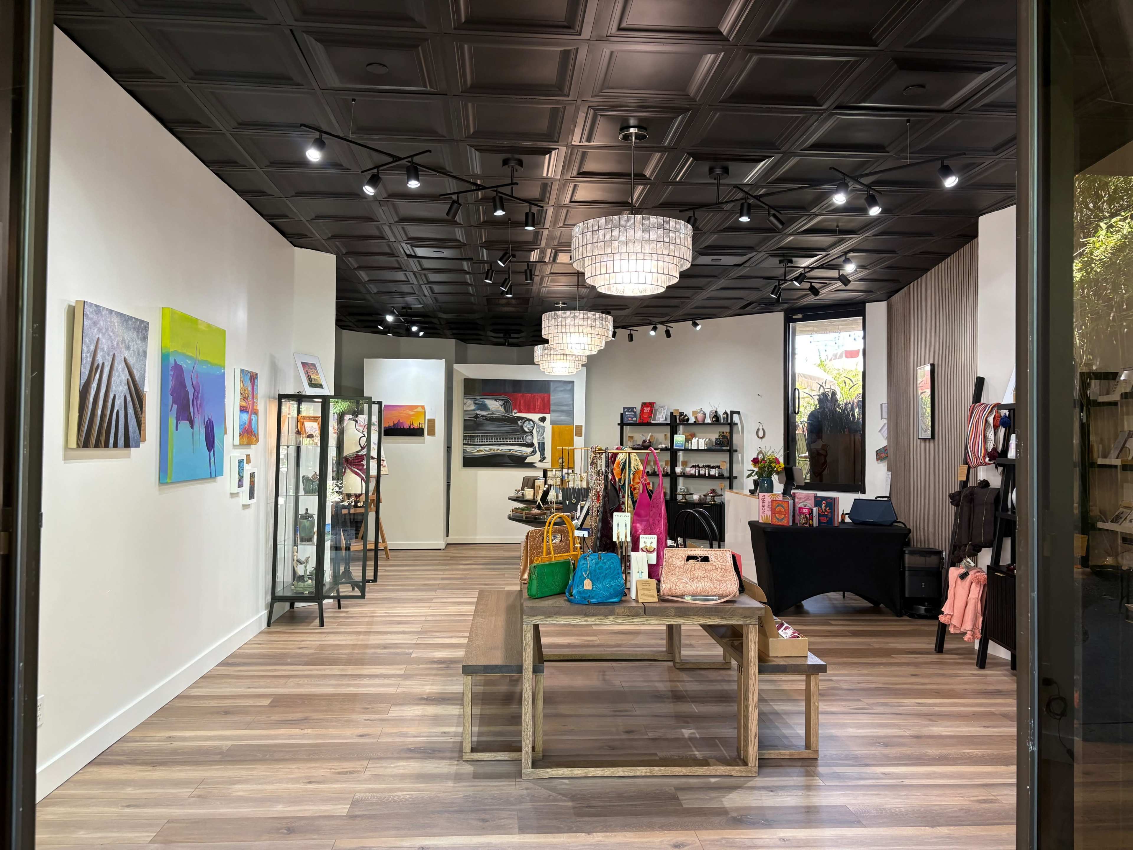 The image shows an art and accessory shop featuring various colorful artworks on the walls, display cases, and shelves with bags and other merchandise arranged on a wooden table.