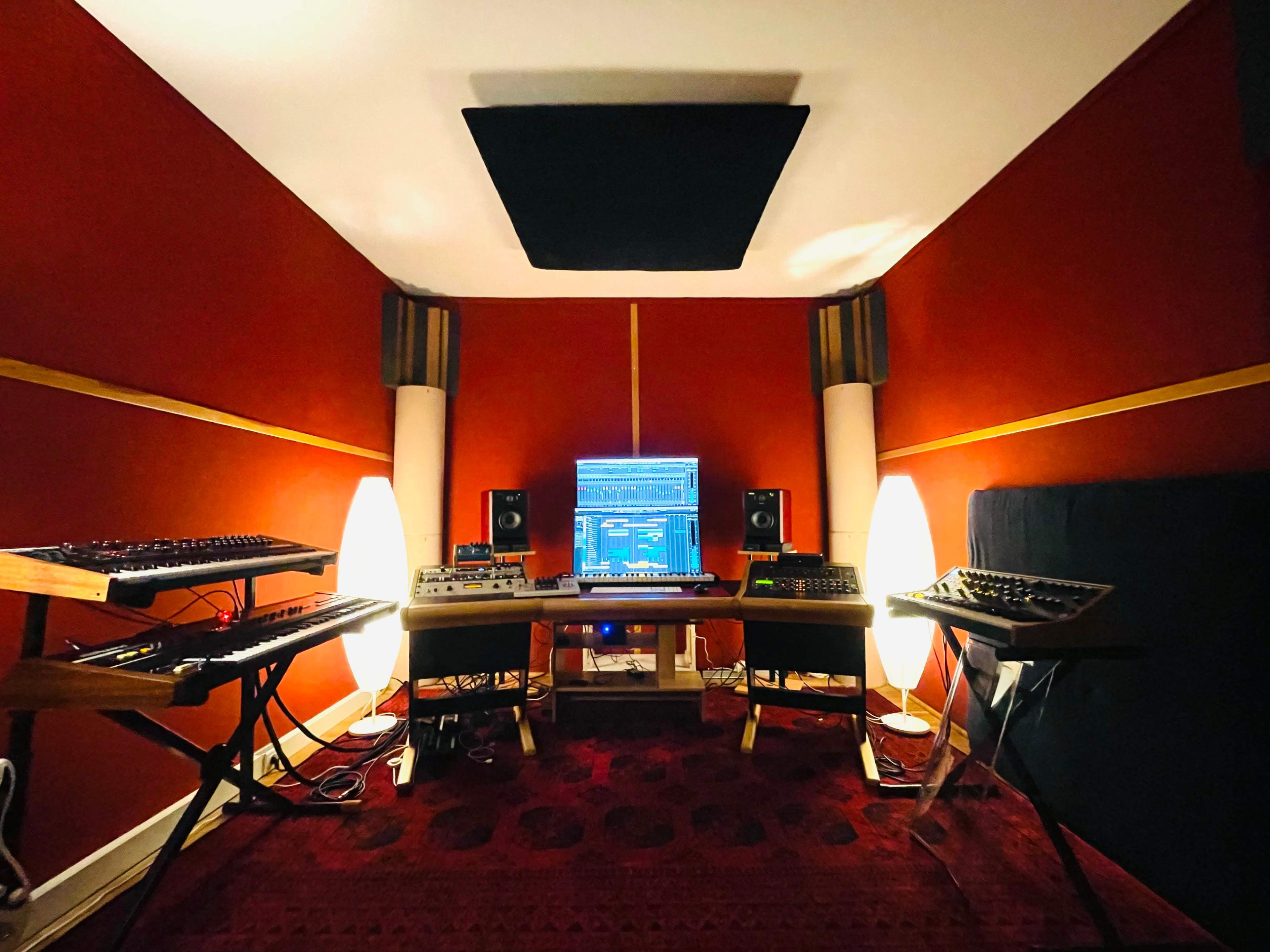 A music production studio with synthesizers and a computer setup against red walls, featuring two illuminated lamps and soundproofing panels.