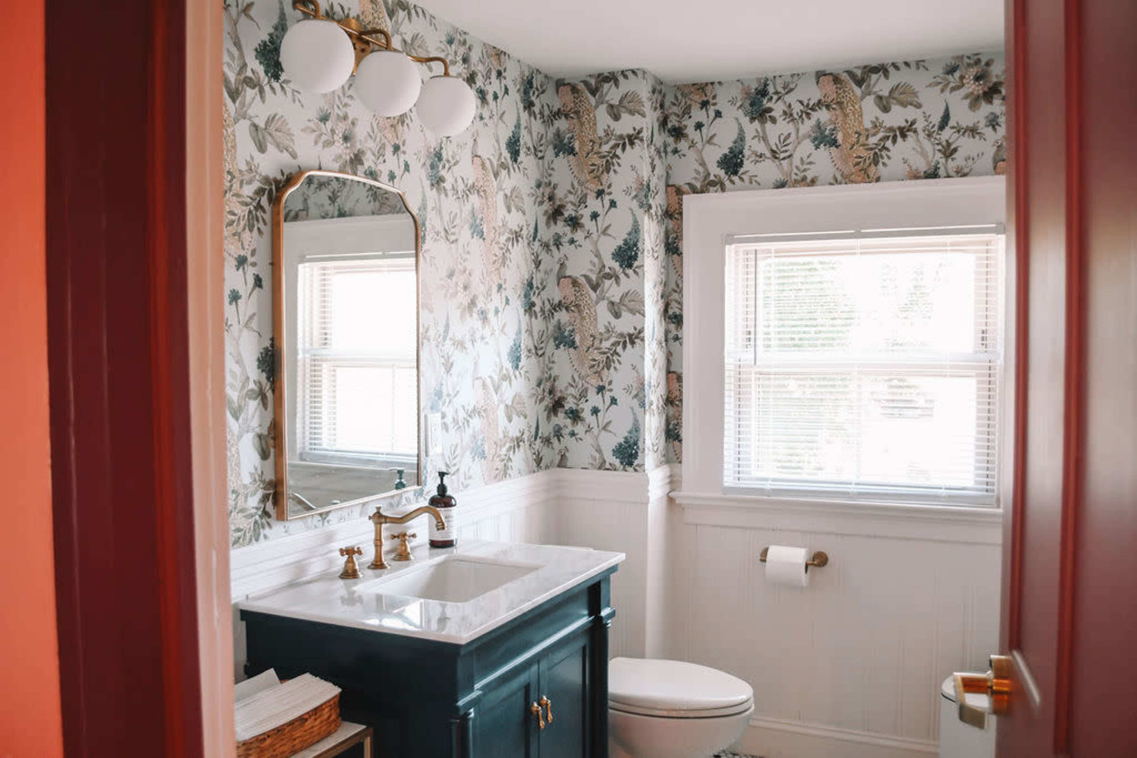 The bathroom features floral-patterned wallpaper, a white countertop with a mirror above, and a toilet beside a window.