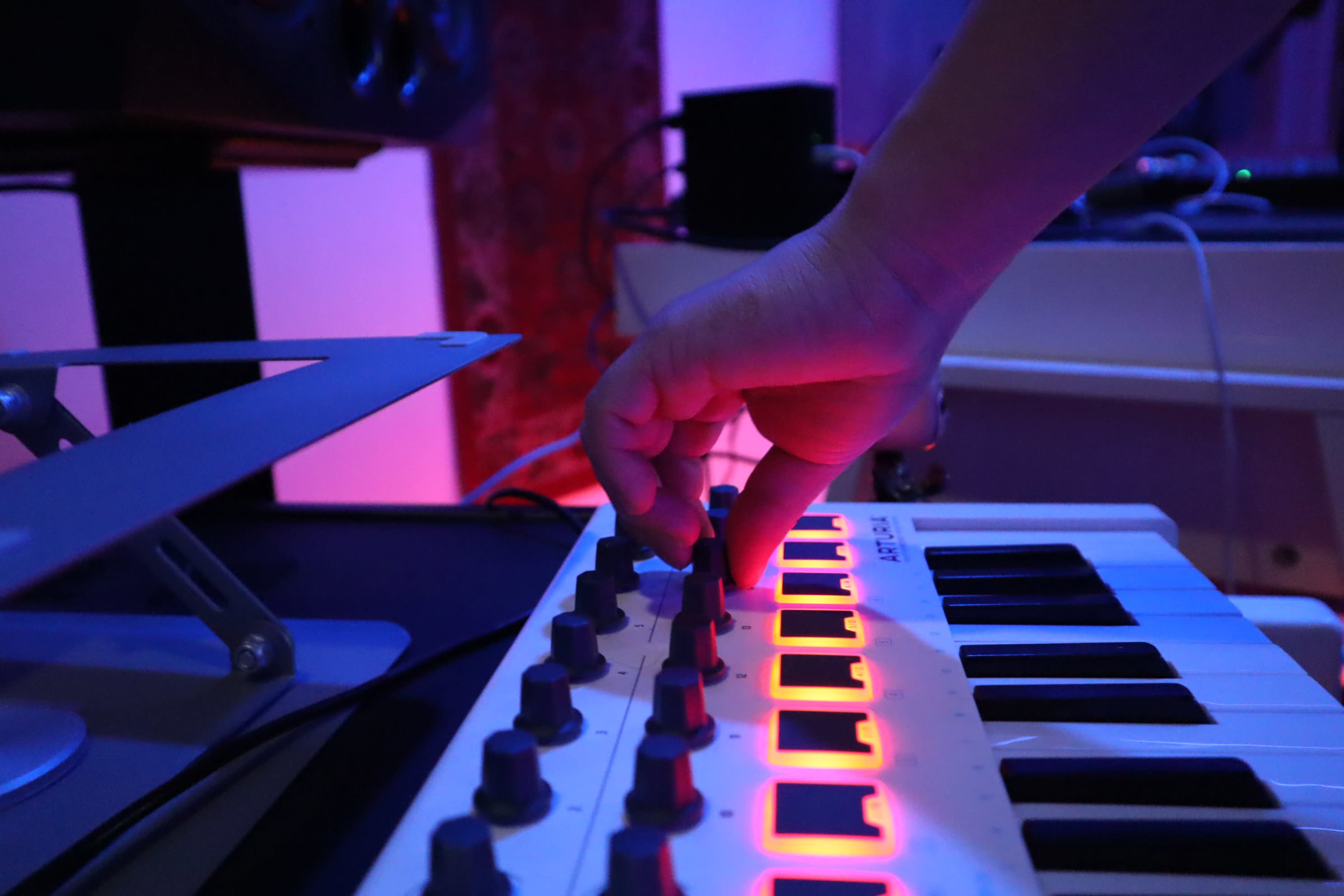 A hand adjusts the knobs on a small synthesizer in a dimly lit studio with multicolored lighting.