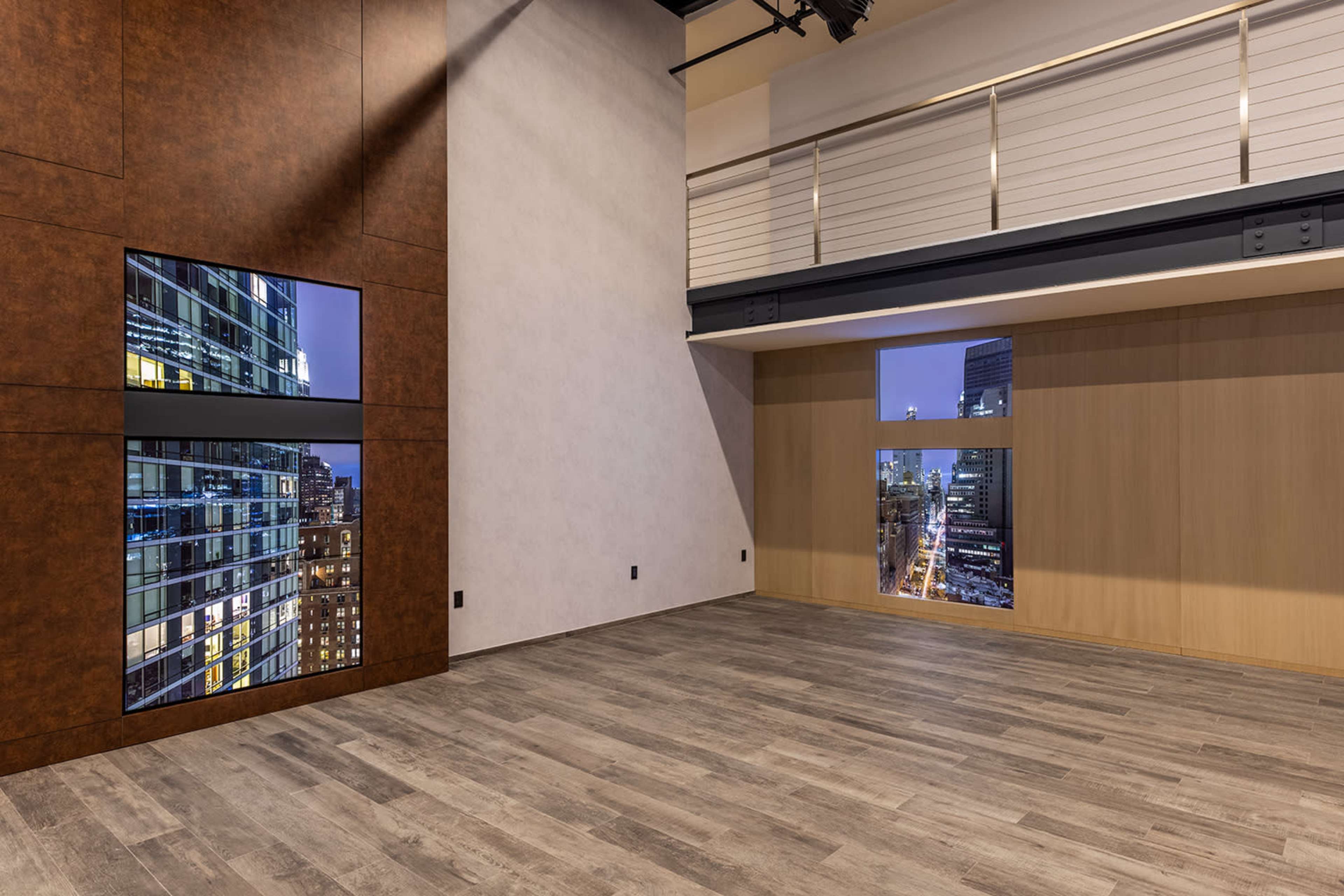 The image shows a modern interior room with large windows revealing a city skyline at night, featuring wooden flooring and a minimalist design.
