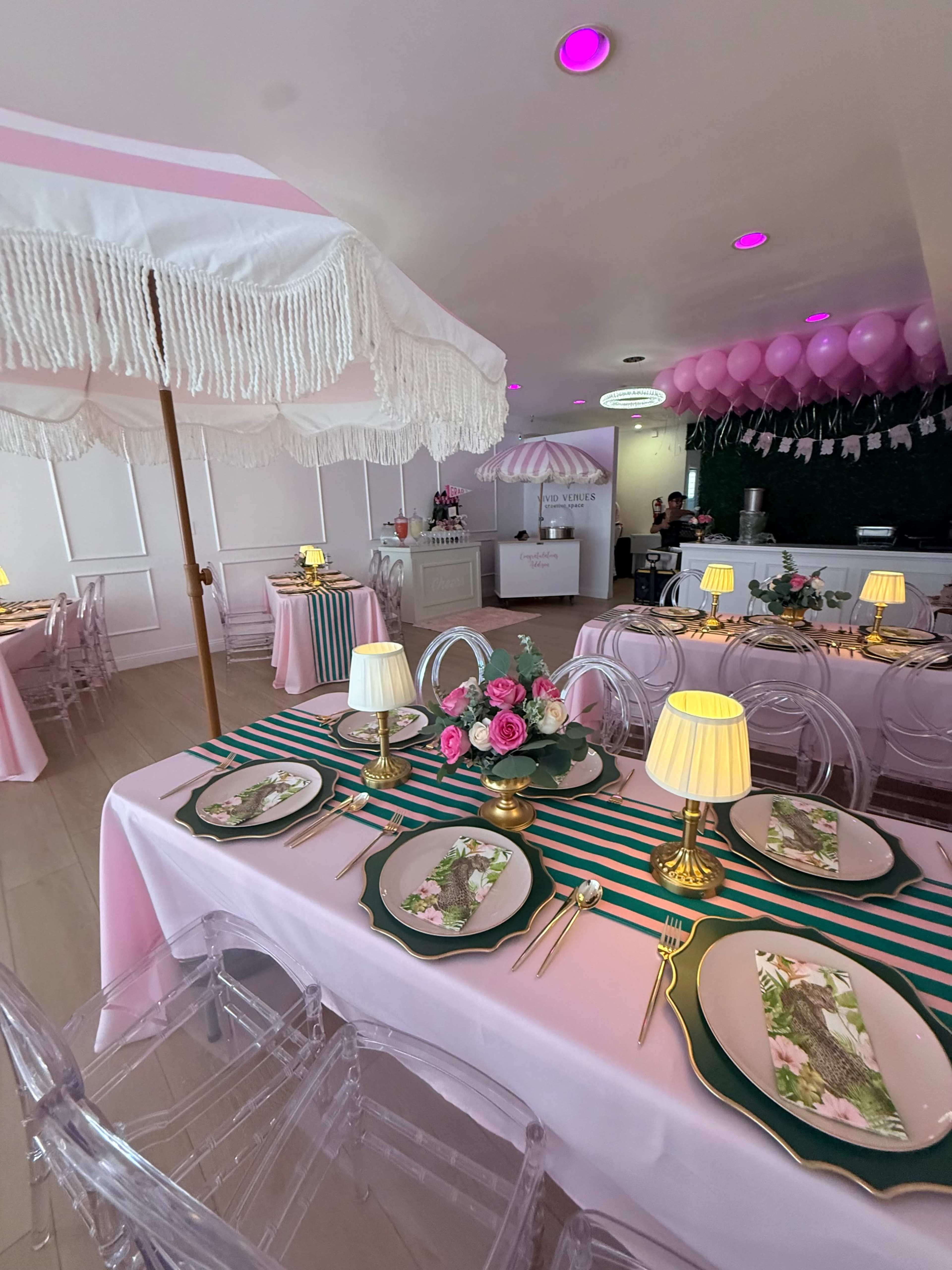 The image shows a brightly decorated café with pastel-colored tables set for dining, featuring floral plates, golden cutlery, and elegant lighting under a canopy.