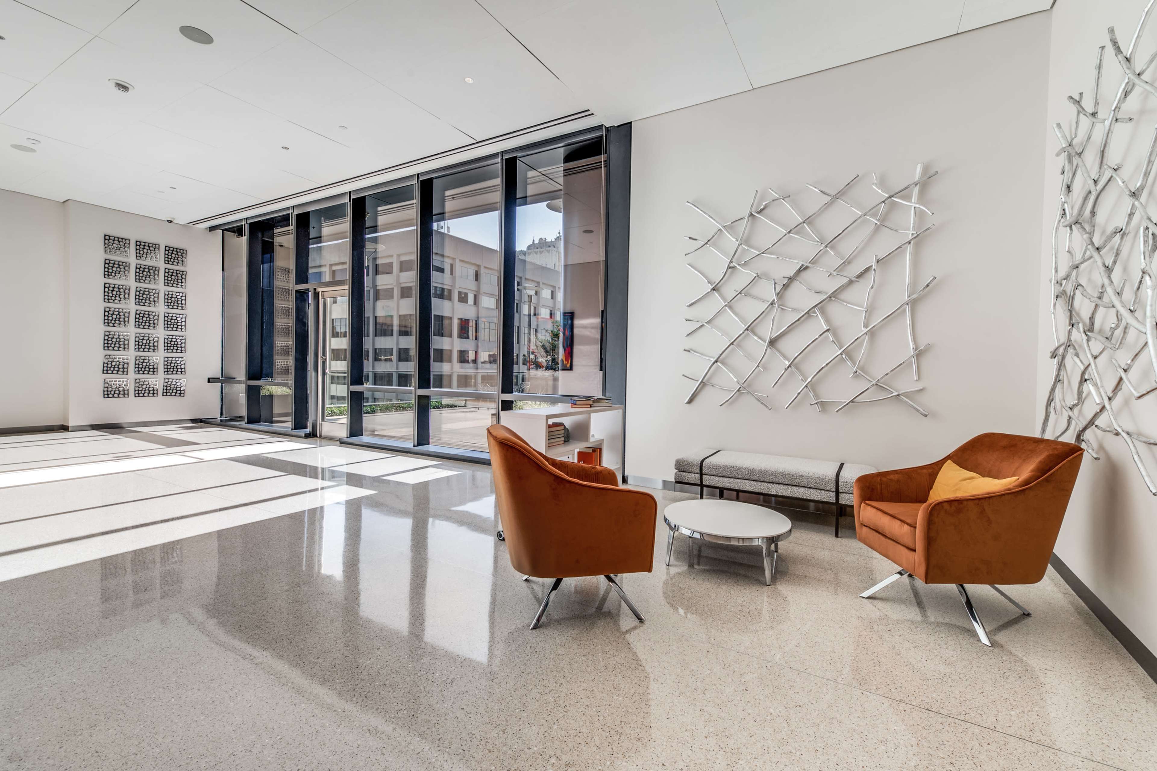 A modern lounge area features two orange armchairs, a round coffee table, and a large window with city views, illuminated by natural light.