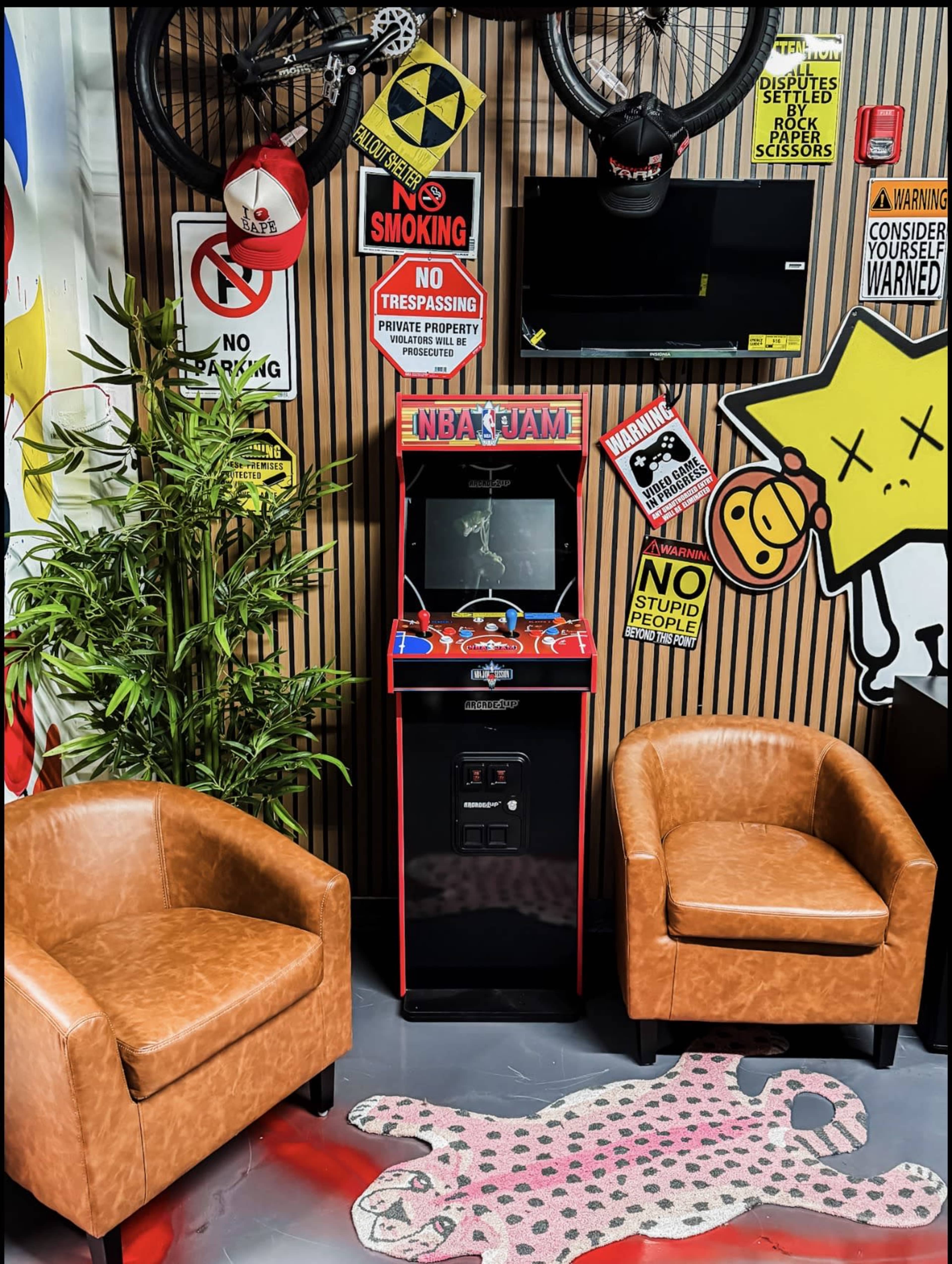 The image features a nostalgic arcade game cabinet for "NBA Jam" positioned between two brown chairs, decorated with various warning signs and a bicycle above in a vibrant game room setting.