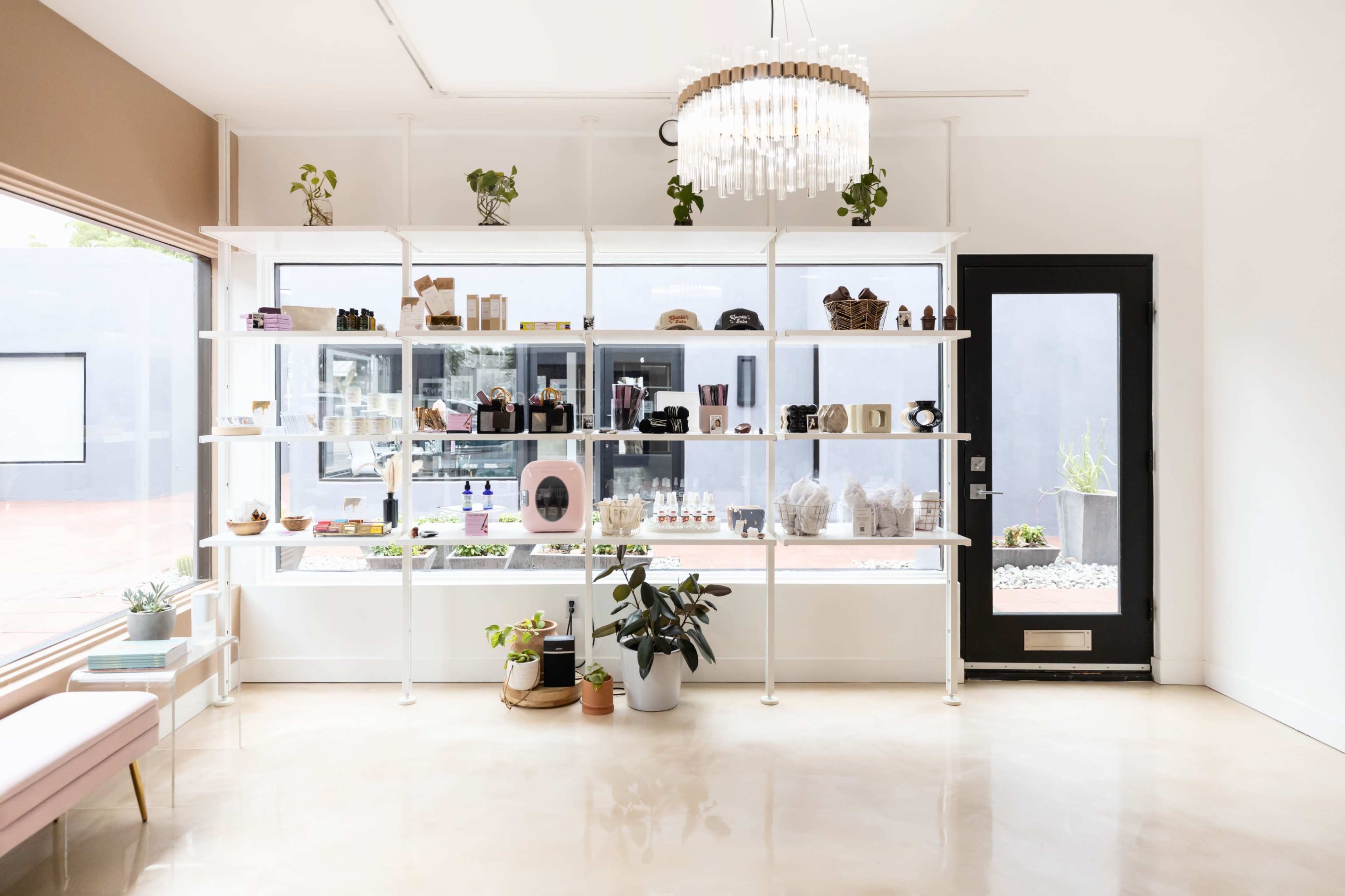 A modern boutique shop with white shelving displaying various products under a chandelier, alongside a large glass door.