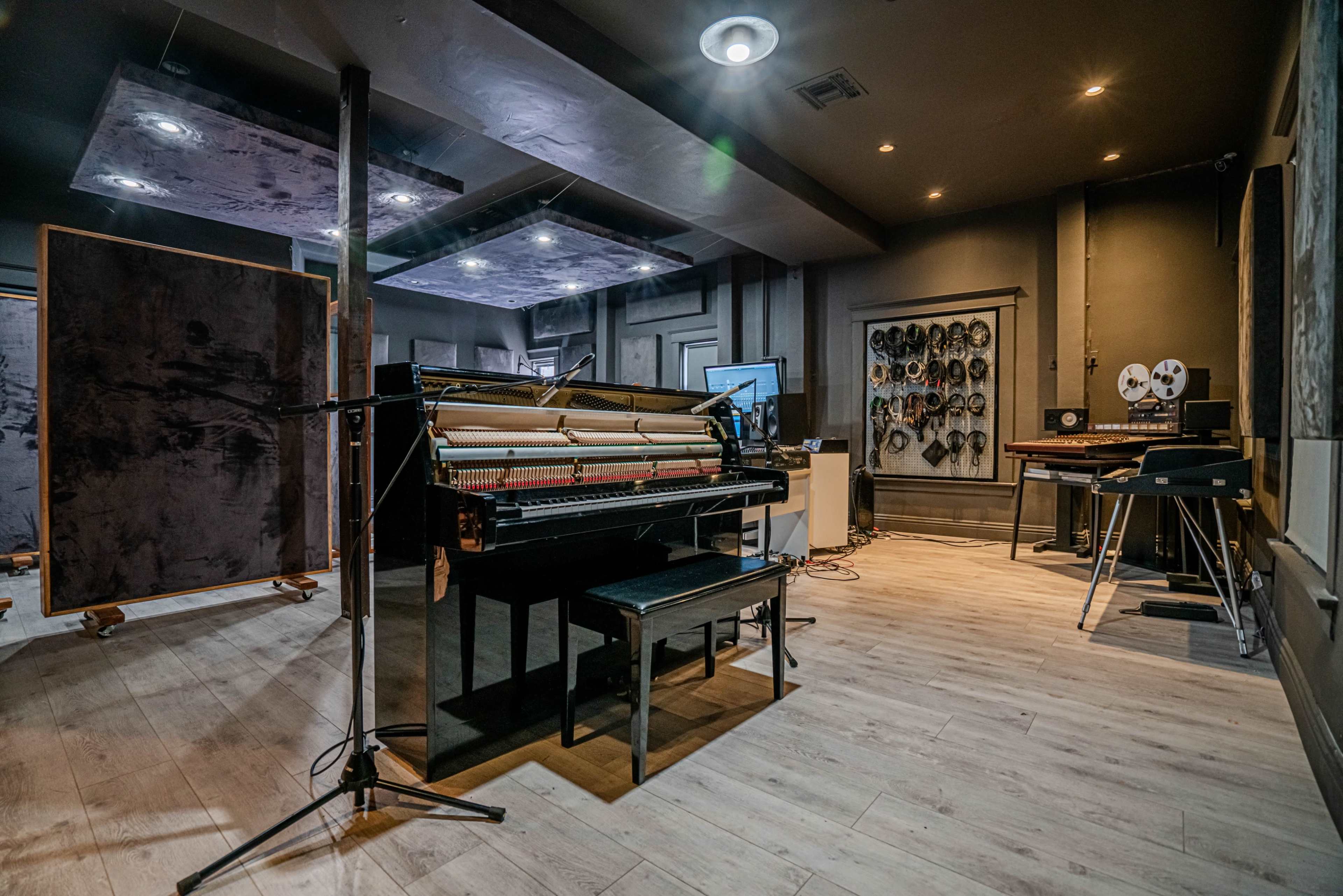 The image shows a recording studio with a black piano at the center, microphones set up, and various musical equipment arranged around the room.