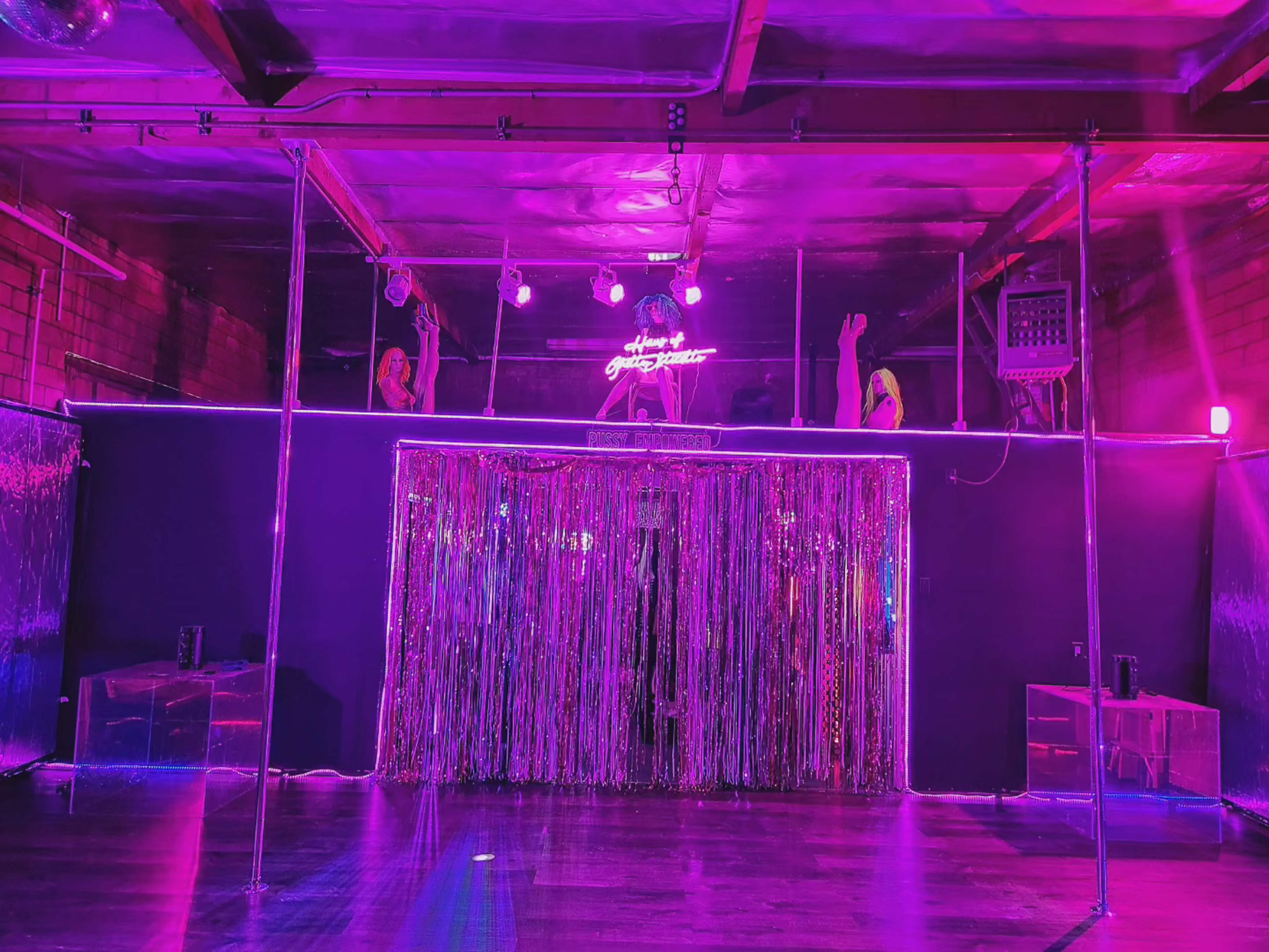 The image shows a dimly lit dance area with a glittering curtain, neon lights, and a stage featuring two poles.