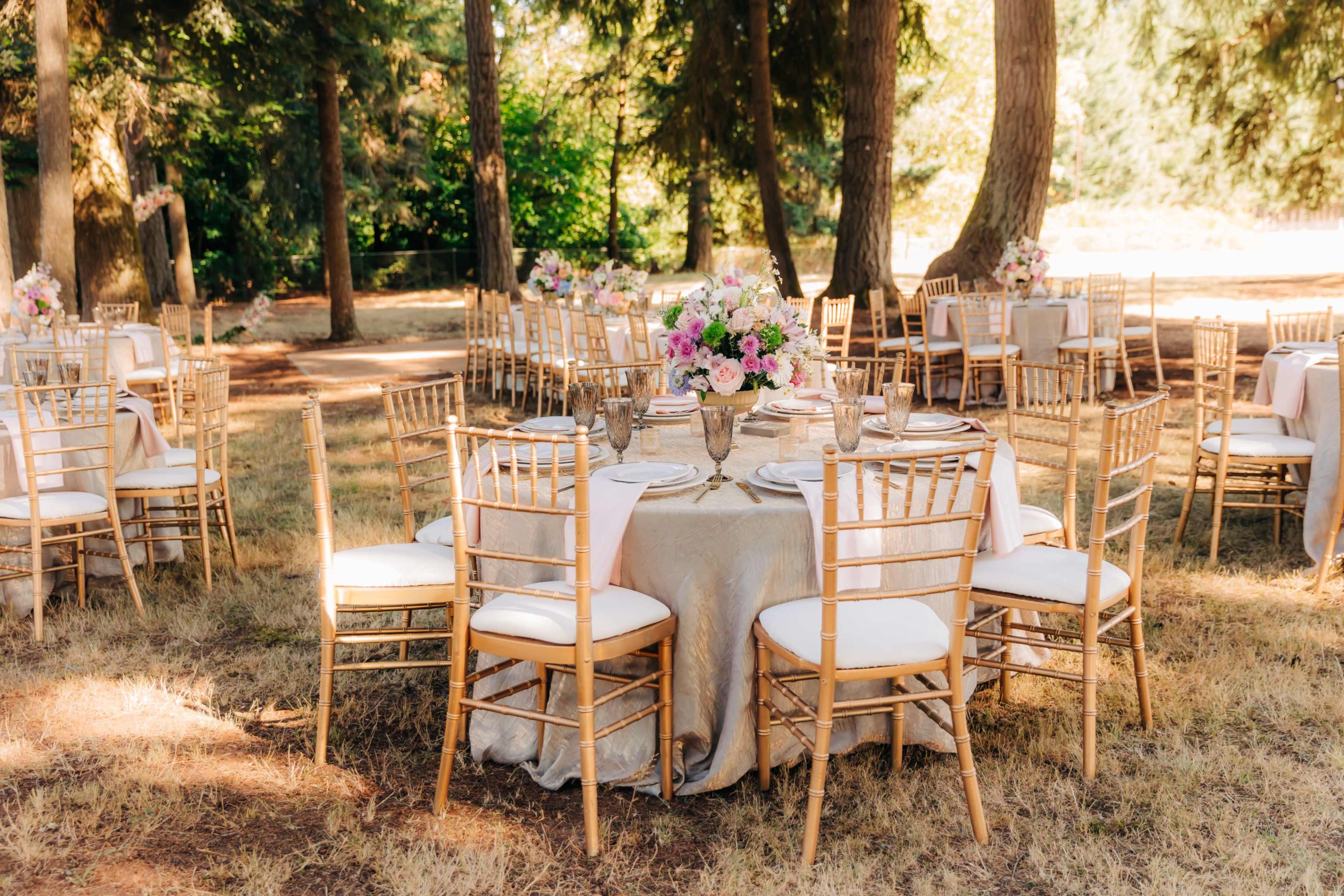 A dining setup beneath tall trees features several round tables adorned with floral centerpieces and golden chairs.