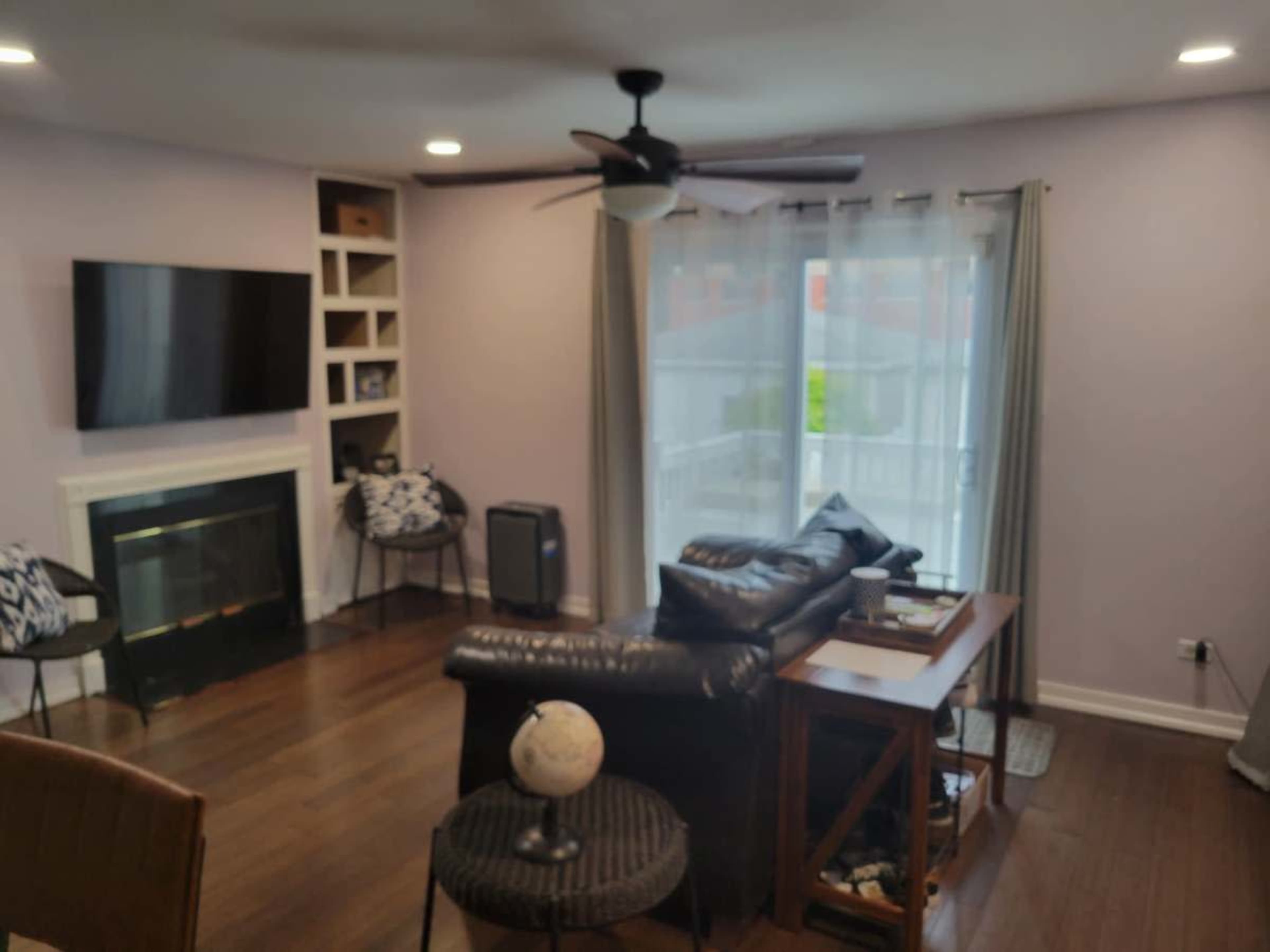 The image shows a living room with a dark leather couch, a wooden side table, a television mounted on the wall, and a window with sheer curtains.