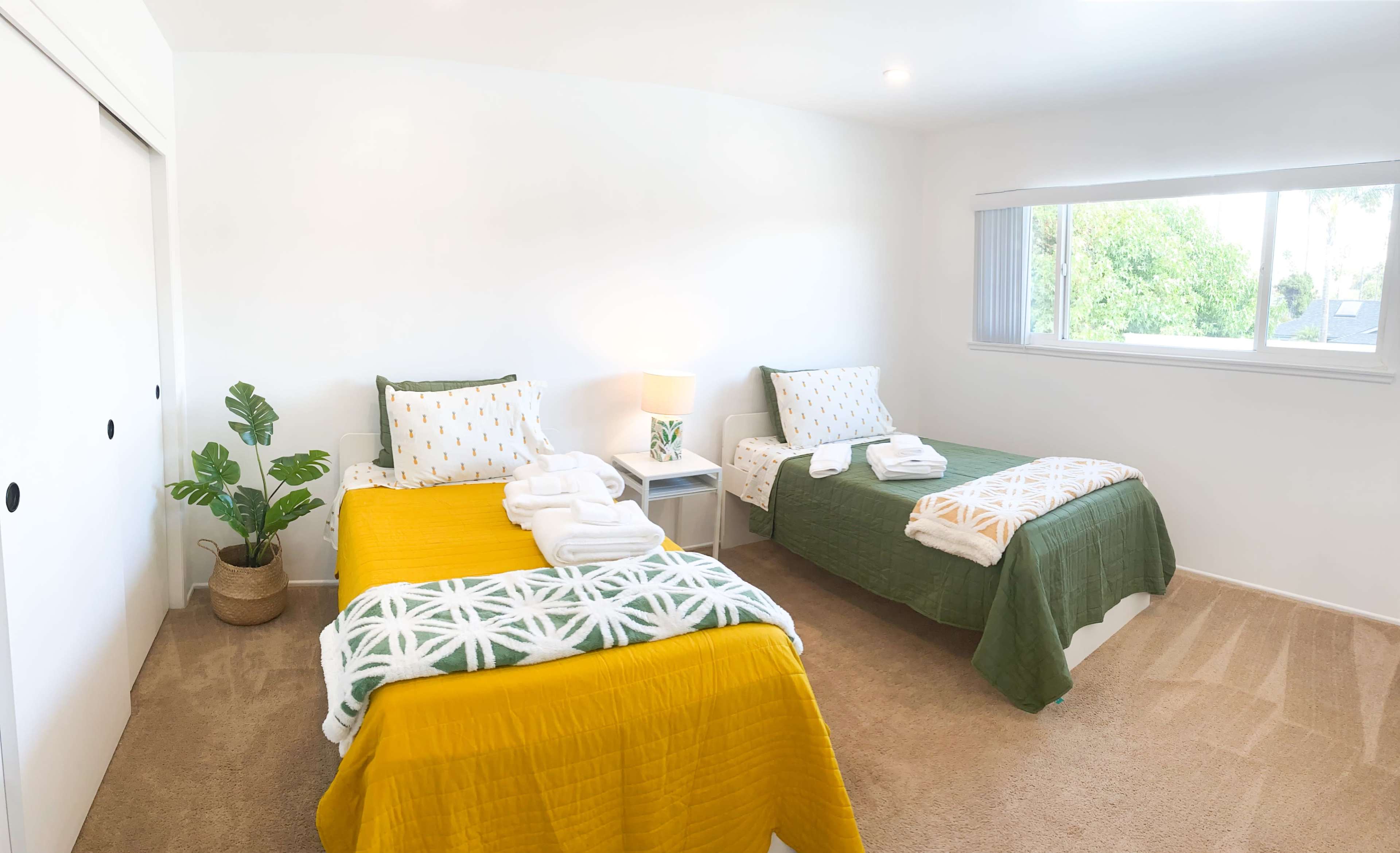 A brightly lit room features two twin beds with yellow and green bedspreads, a nightstand with a lamp, and a potted plant by the window.