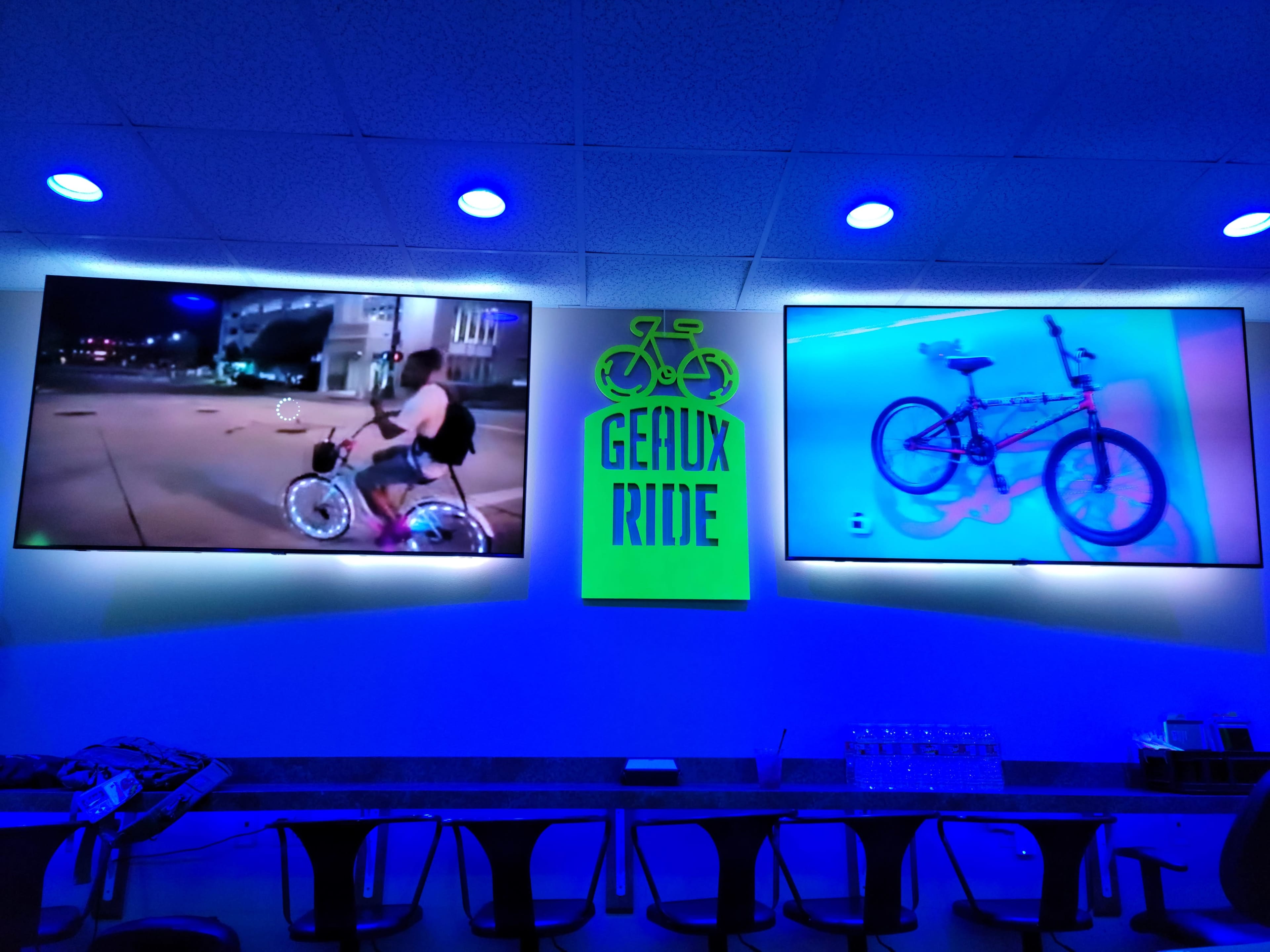 Two televisions mounted on a wall, displaying videos of biking; a neon sign in the center reads "Geraux Ride."
