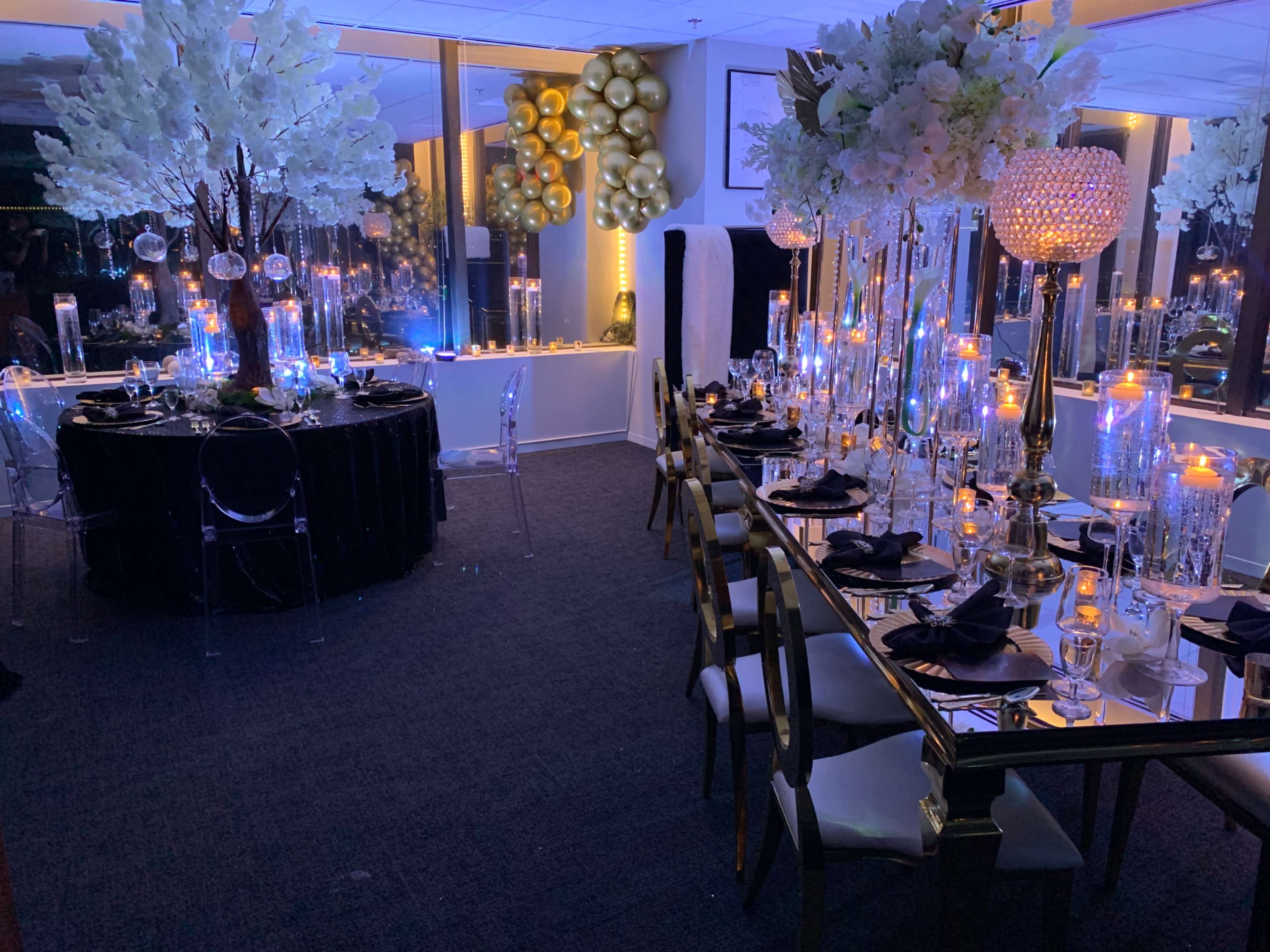 An elegantly decorated event space with tables set for dining, featuring tall floral arrangements, candles, and balloons.