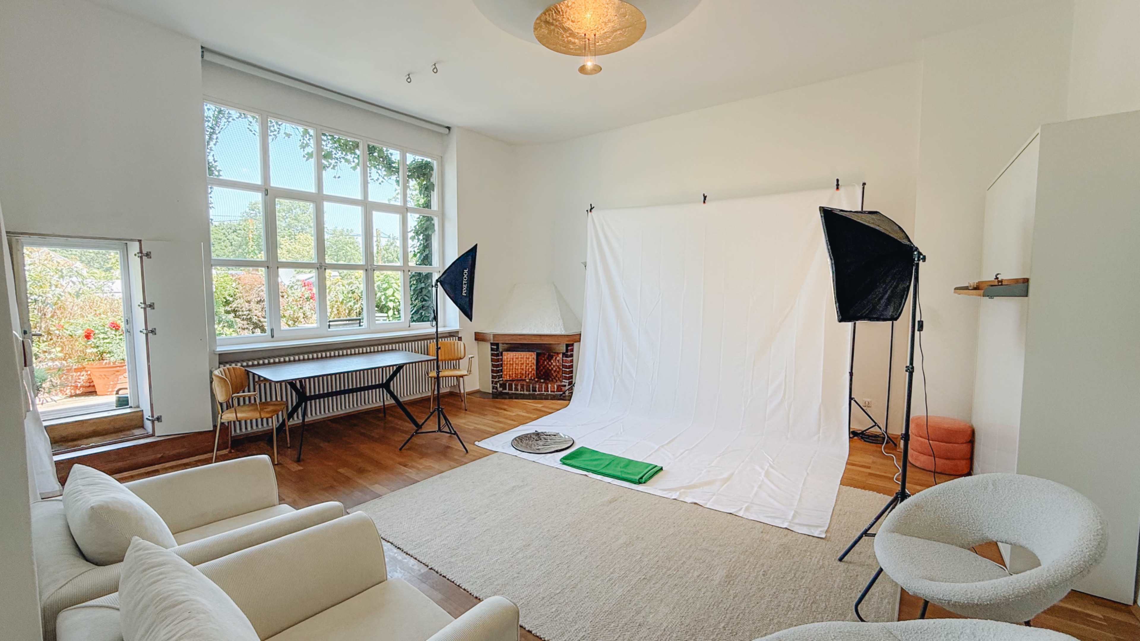 The image shows a photography studio with a large window, white backdrop, lighting equipment, and simple furniture.