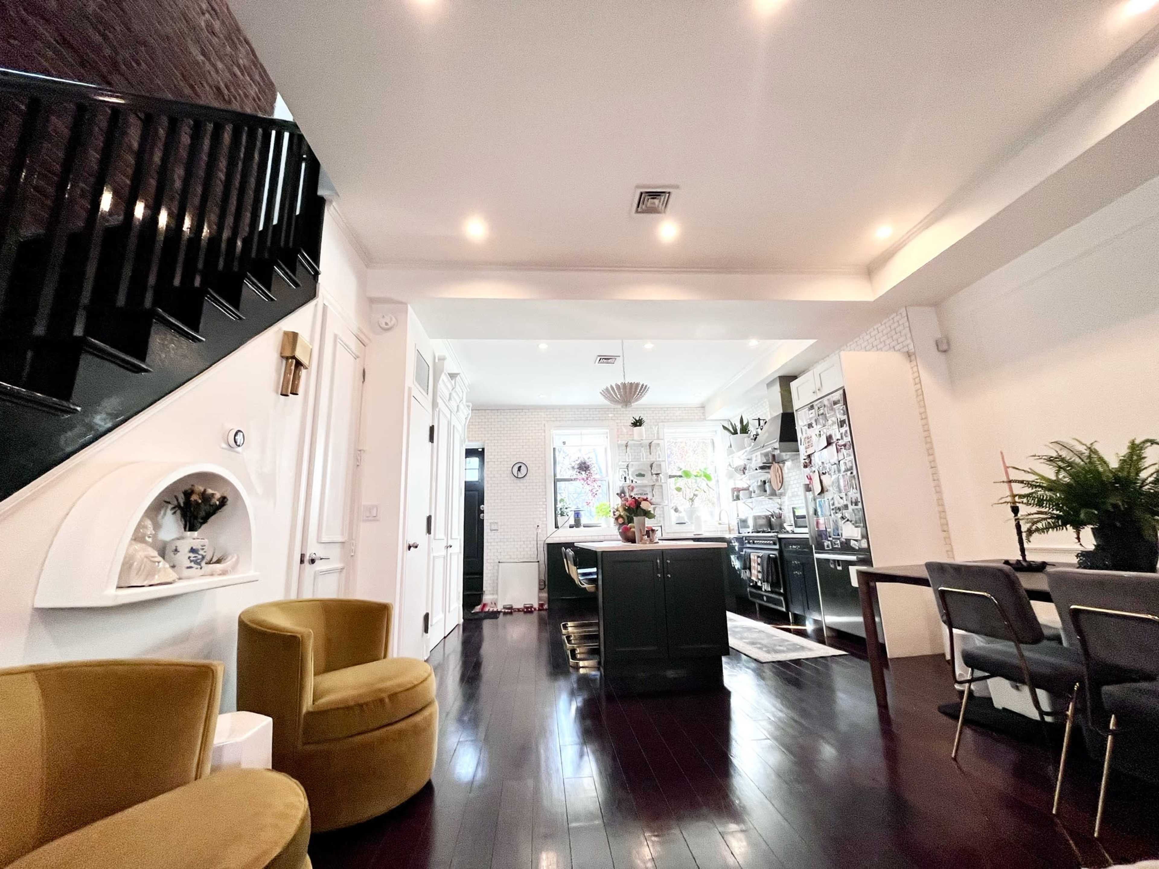 The image shows a modern kitchen and dining area with a staircase, featuring dark wood flooring, green cabinets, and a dining table with gray chairs.