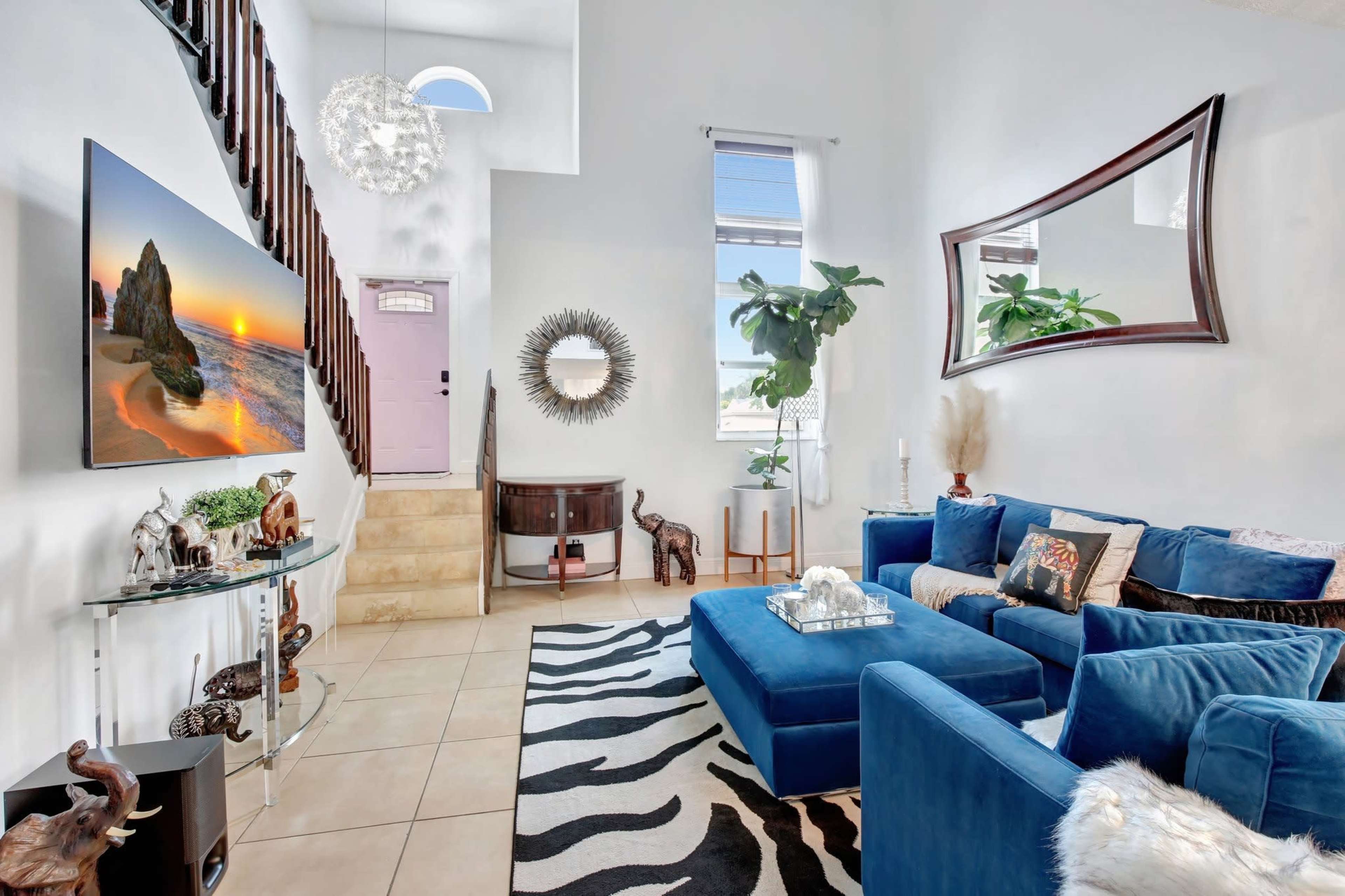 The interior of a modern living room features a blue sectional sofa, a large wall-mounted television, decorative mirrors, and a staircase leading to an upper level.