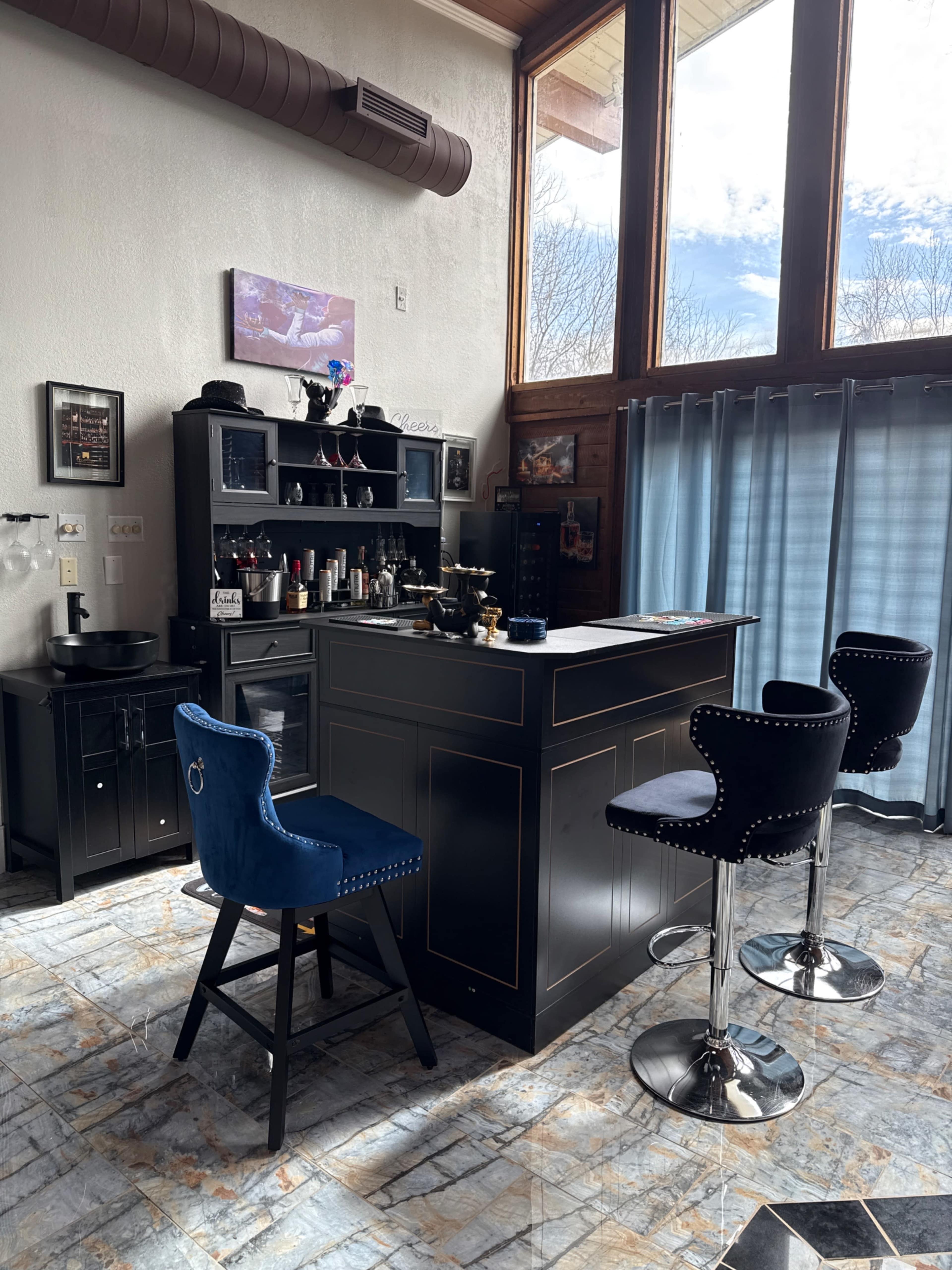 The image shows a modern home bar with a black counter, a shelving unit filled with various barware, and three stylish bar stools.