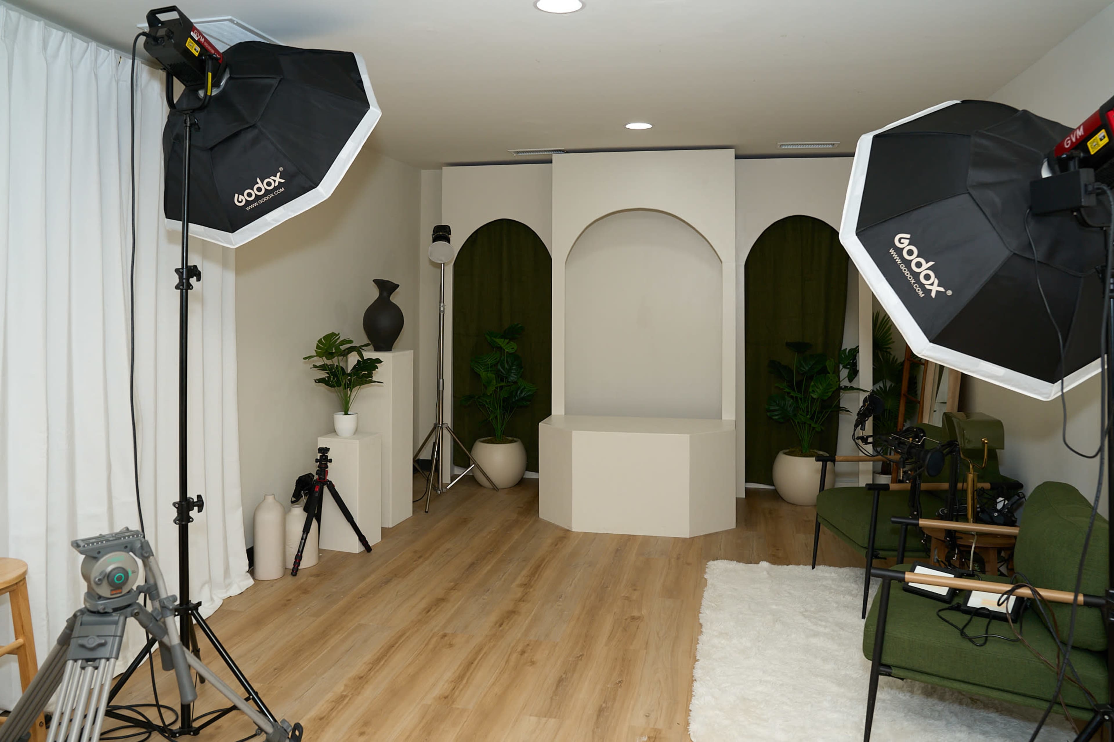 The image shows a photography studio setup with two softbox lights, a simple backdrop featuring an arch, and green plants positioned in the corners.