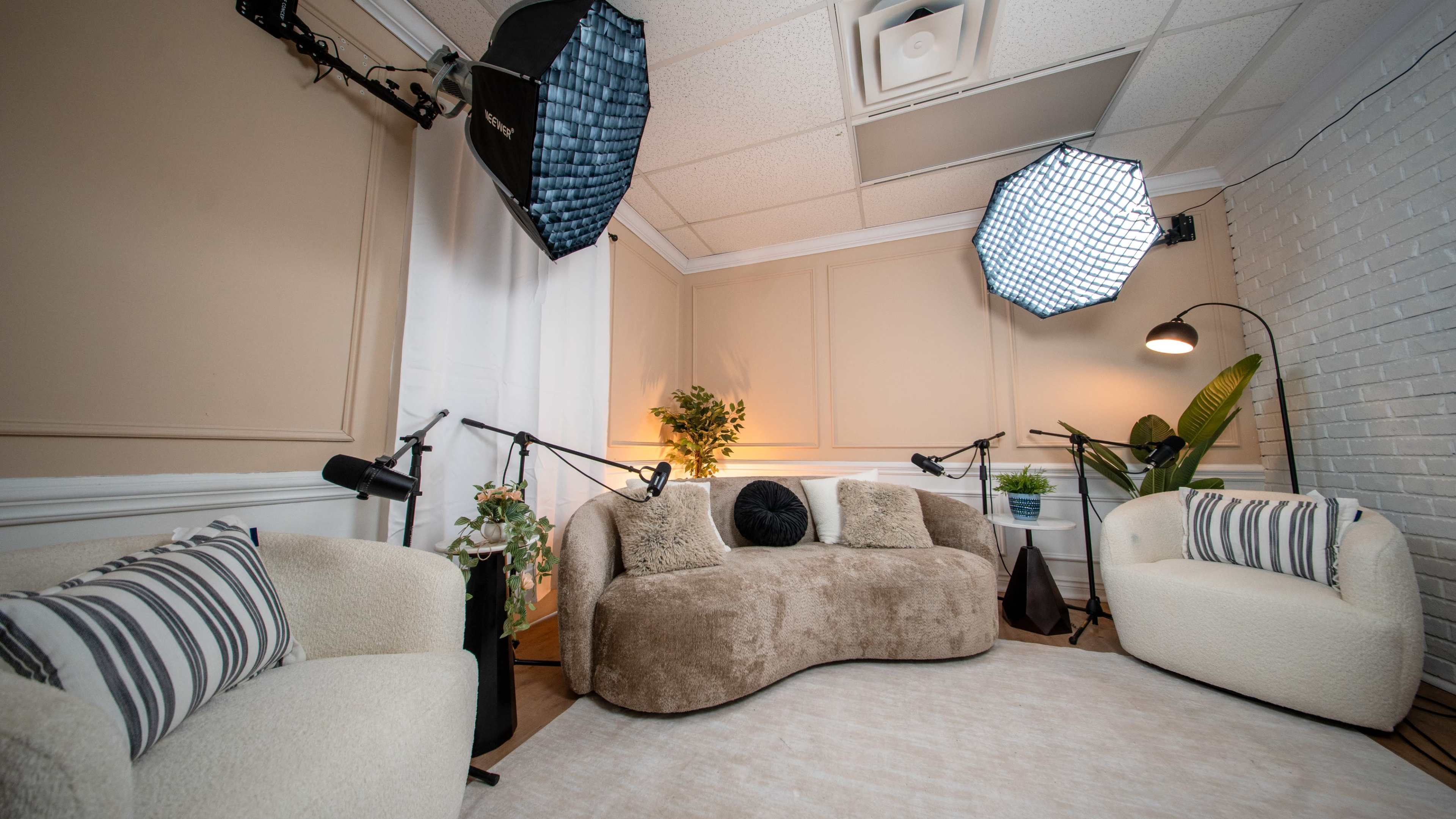 The image shows a well-lit studio setup featuring two curved couches, two armchairs, and various lighting equipment against a backdrop of beige walls and a white brick partition.