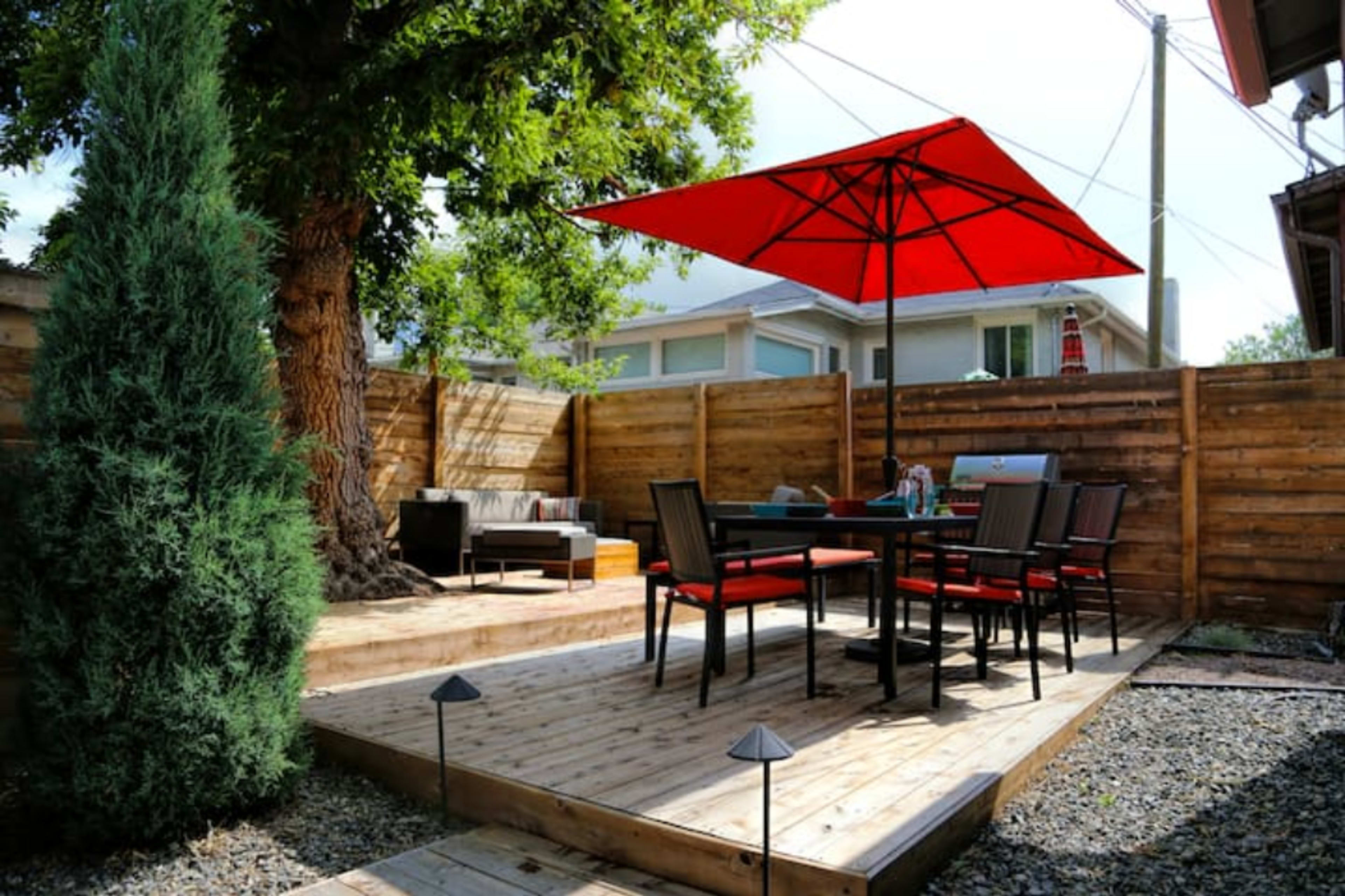 A wooden patio features a table and chairs under a red umbrella, surrounded by wooden fencing and greenery.