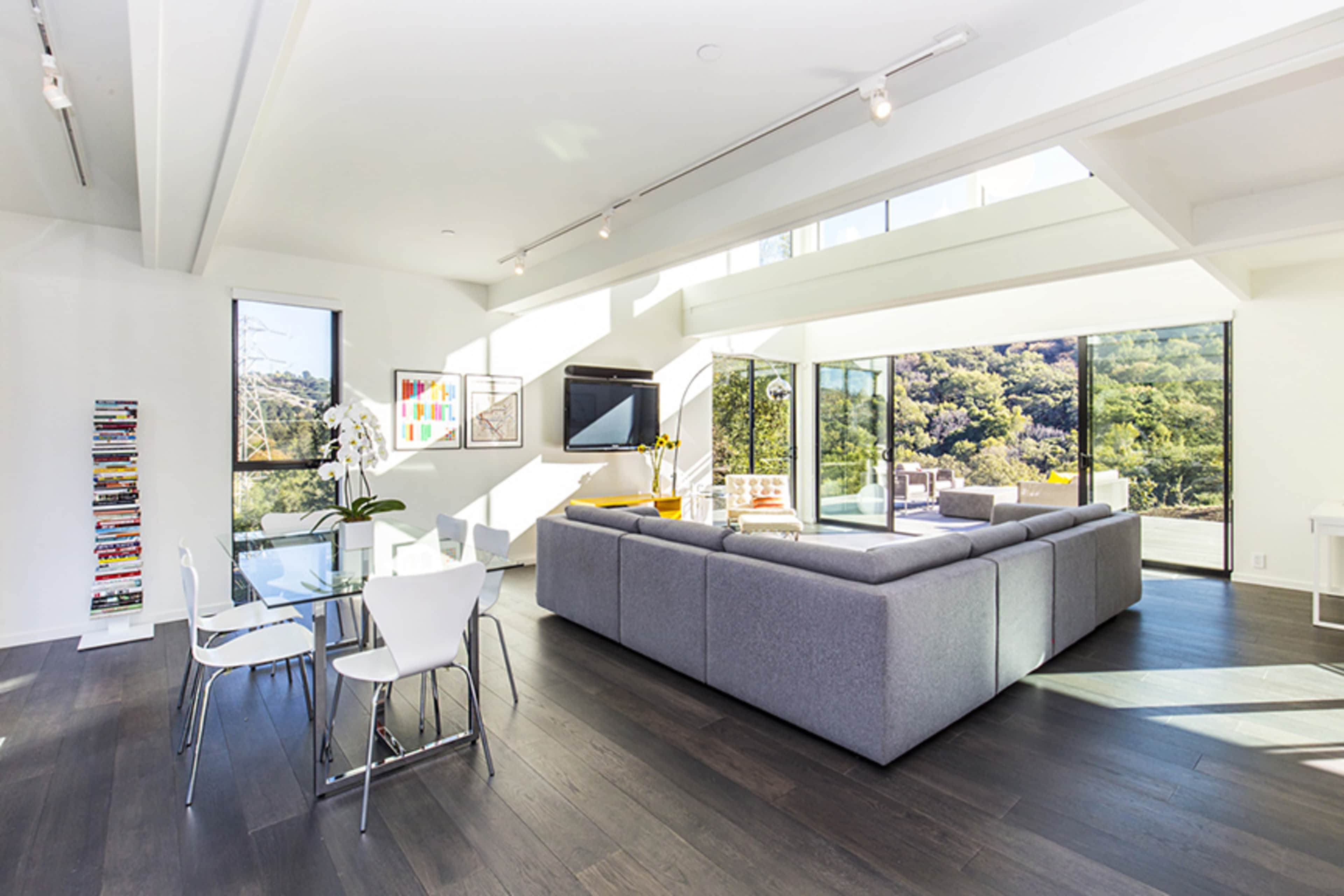A modern living space featuring a gray sectional sofa, a glass dining table with white chairs, and large windows that provide a view of the surrounding hillside.