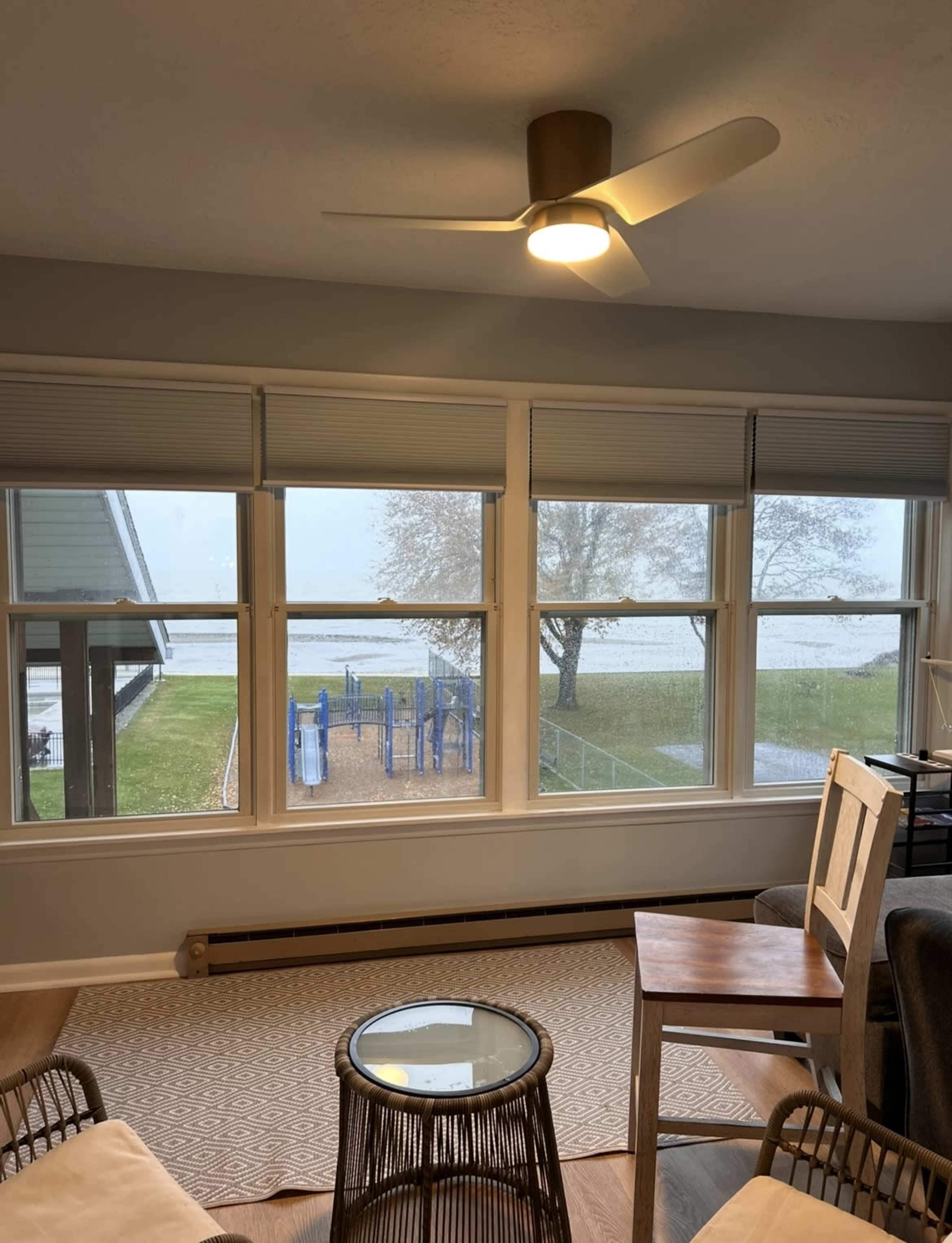 The image shows a cozy indoor space with a ceiling fan, a small table with a glass top, and large windows overlooking a playground and a body of water outside on a rainy day.