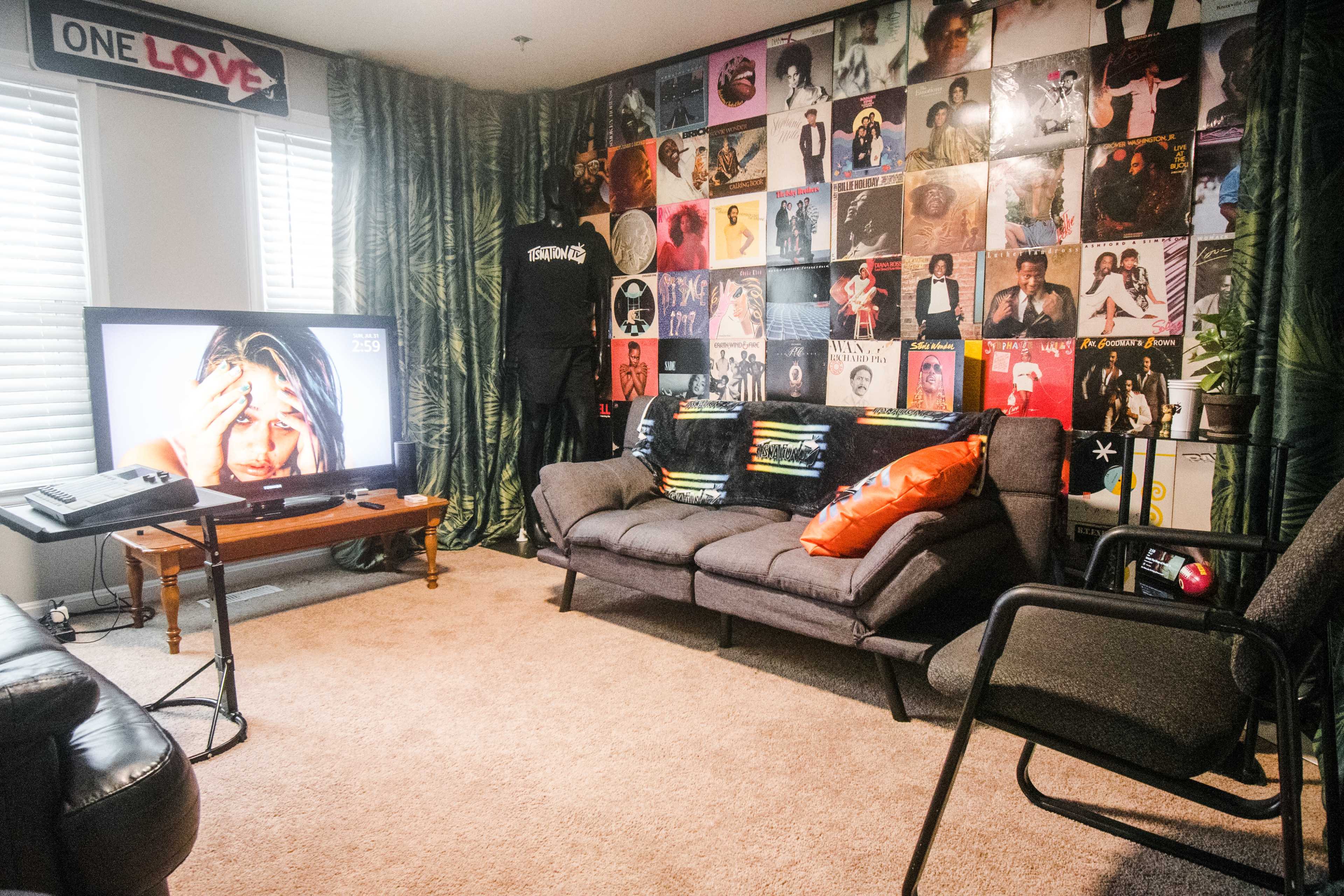 The room features a television displaying a distressed woman and is decorated with a wall covered in vinyl record covers, a gray sofa, colorful cushions, and a small wooden table.