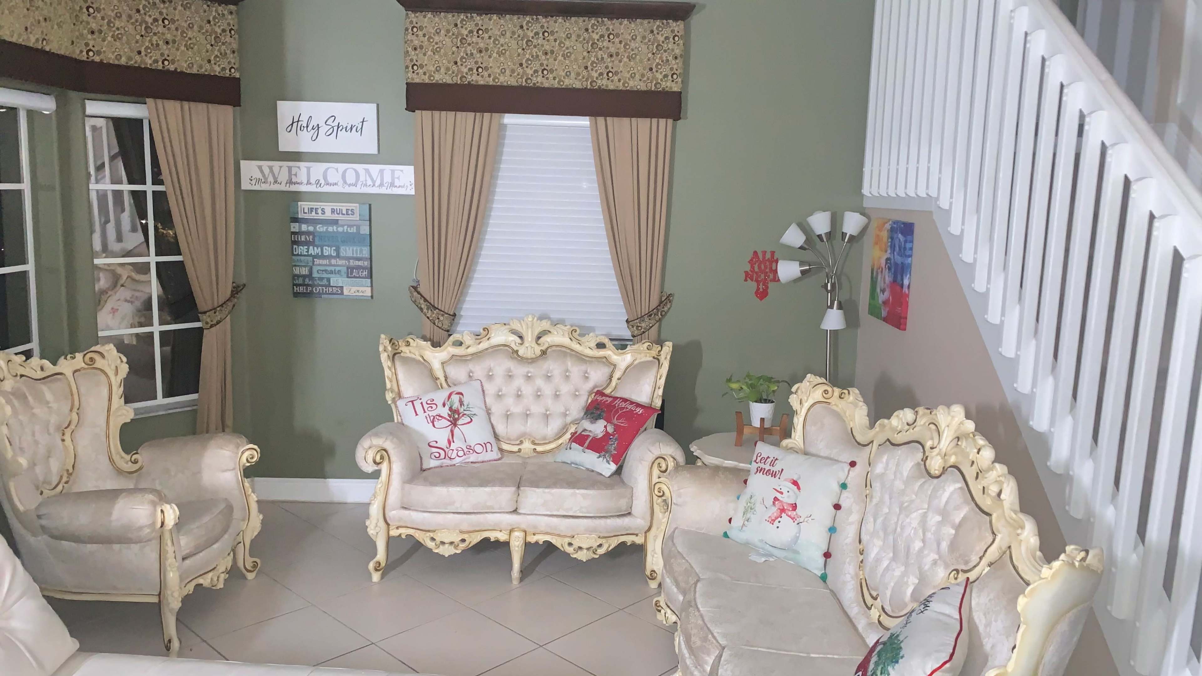 The image shows a living room with ornate light-colored furniture, decorated with festive pillows, and a staircase visible in the background.