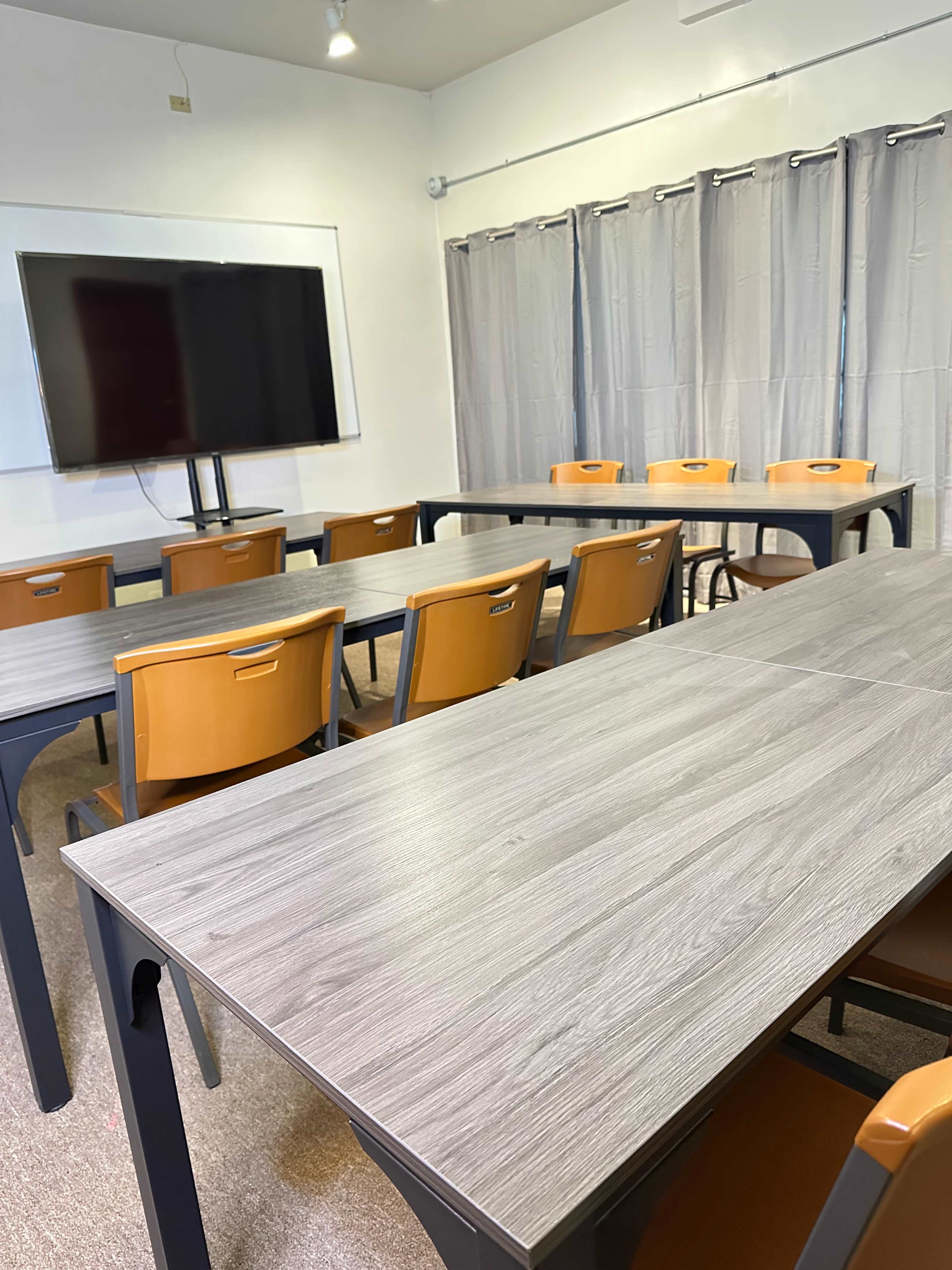 The image shows a classroom with several tables arranged in rows, orange chairs, and a large TV mounted on the wall.