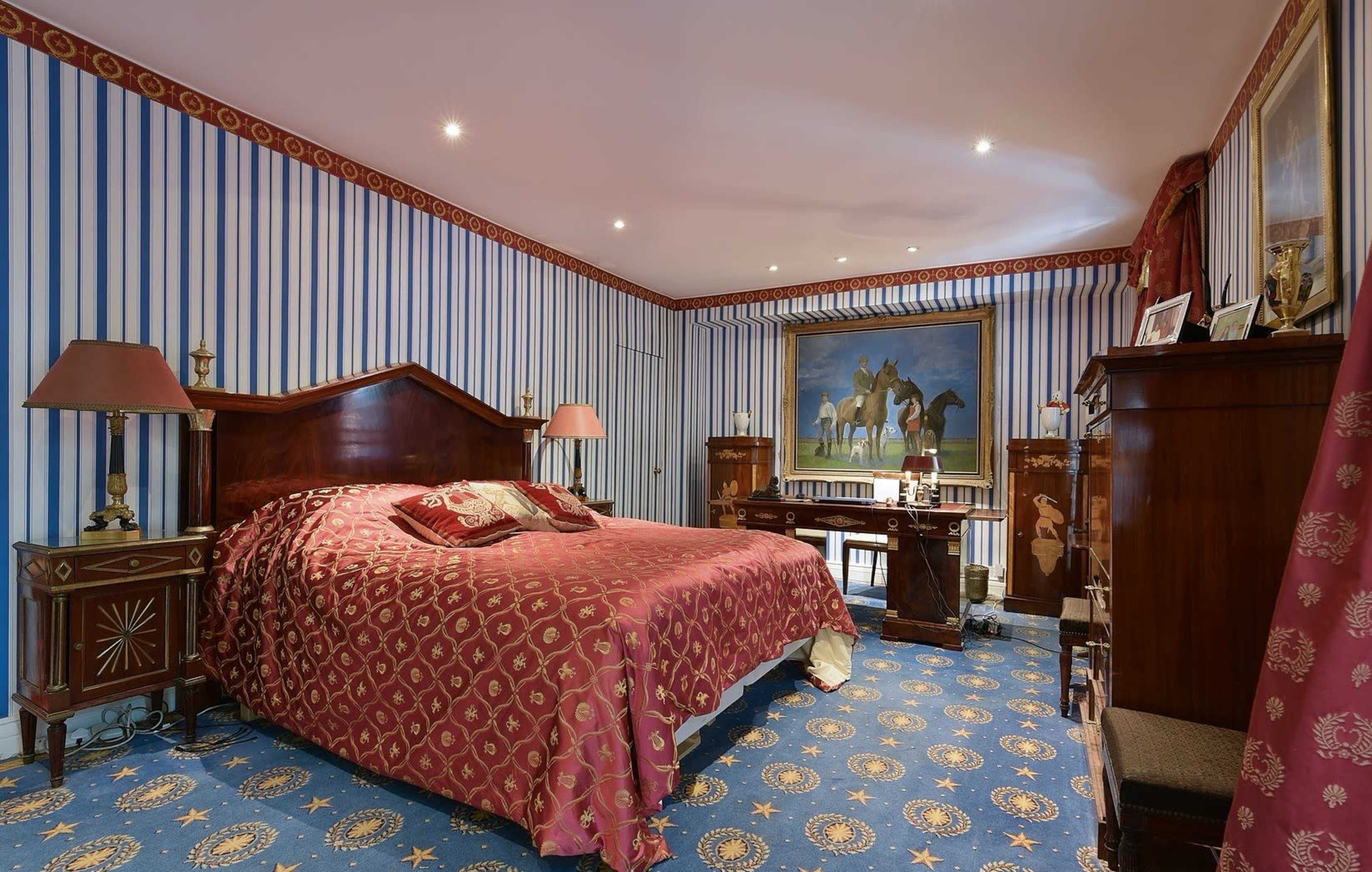 The image shows a bedroom with striped wallpaper, a large bed covered in a red patterned bedspread, and various furniture pieces, including a desk and ornate lamps.