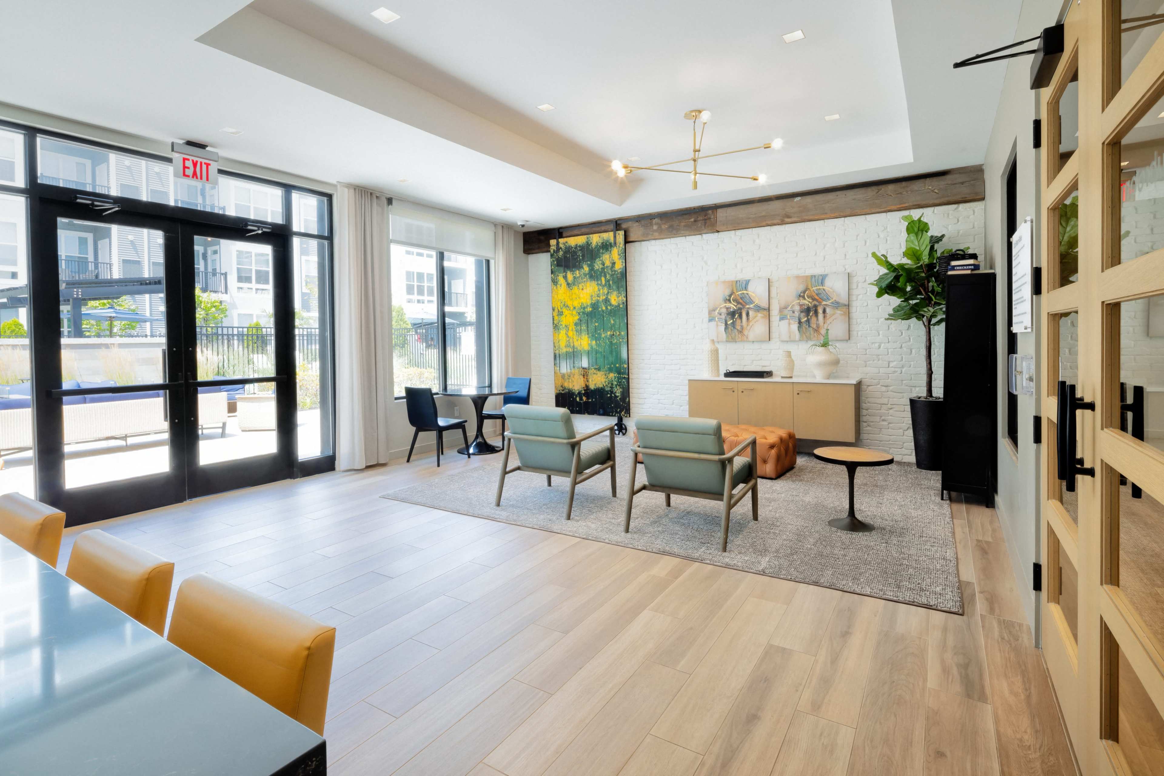A modern lounge area with a seating arrangement, a small table, and a decorative wall art piece, all set against large windows and a light-colored brick wall.
