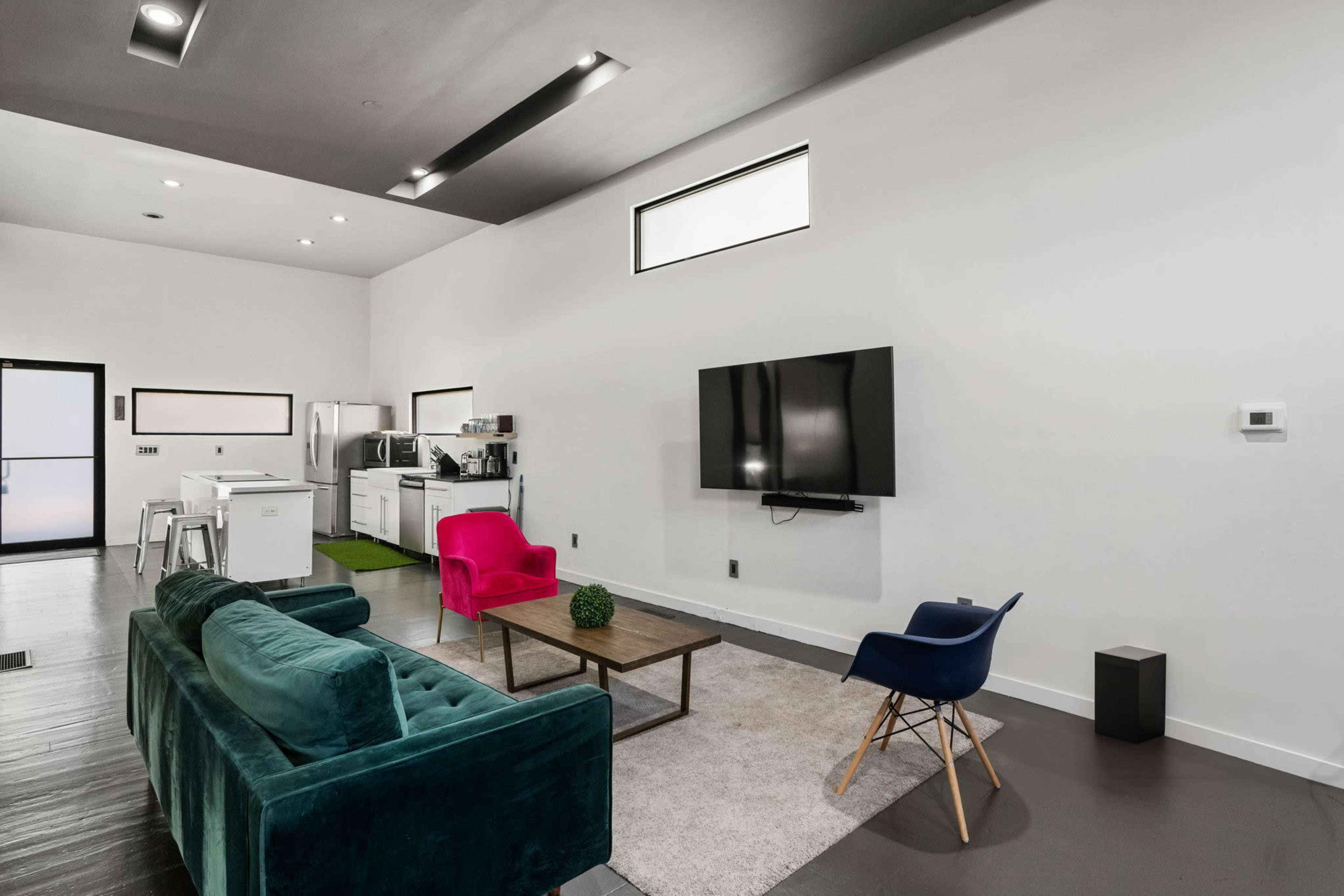 A modern living space with a green velvet sofa, a pink chair, a wooden coffee table, a large flat-screen TV on the wall, and a kitchen area in the background.