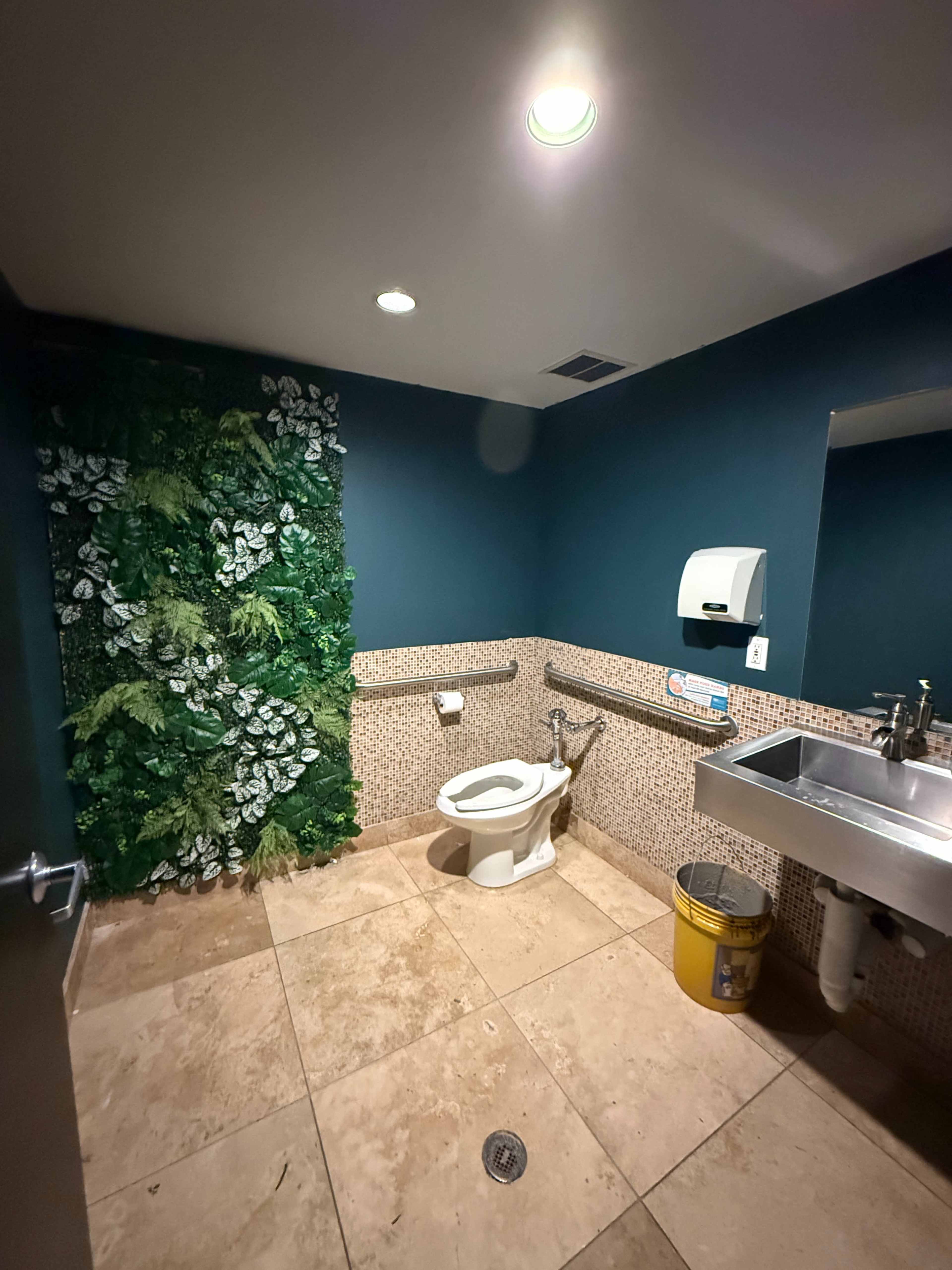 A restroom features a toilet, a wall with greenery, a sink, and tiled flooring.