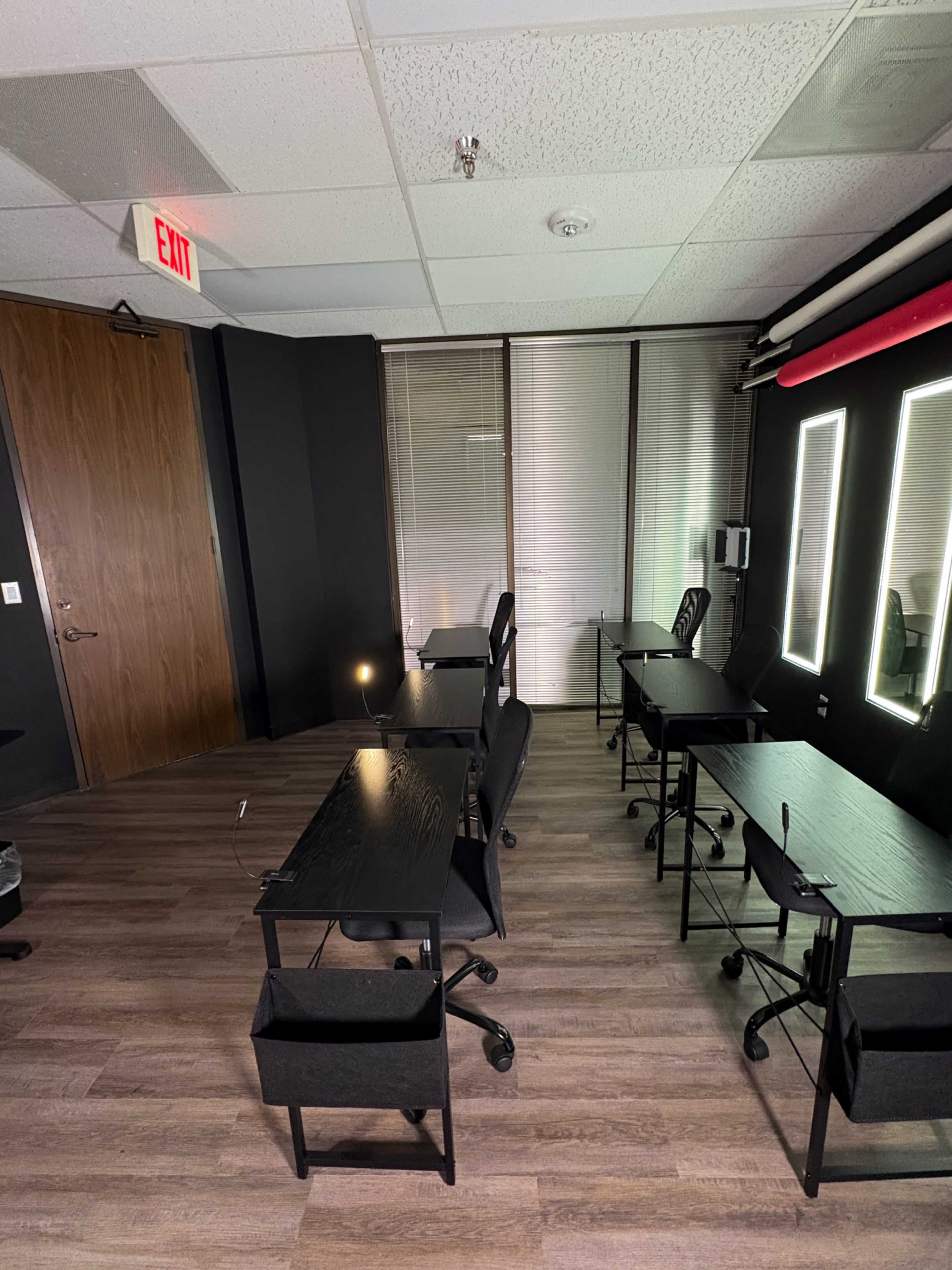 The image shows a small, sparsely furnished room with several black desks and chairs, illuminated by lighted mirrors on one wall.