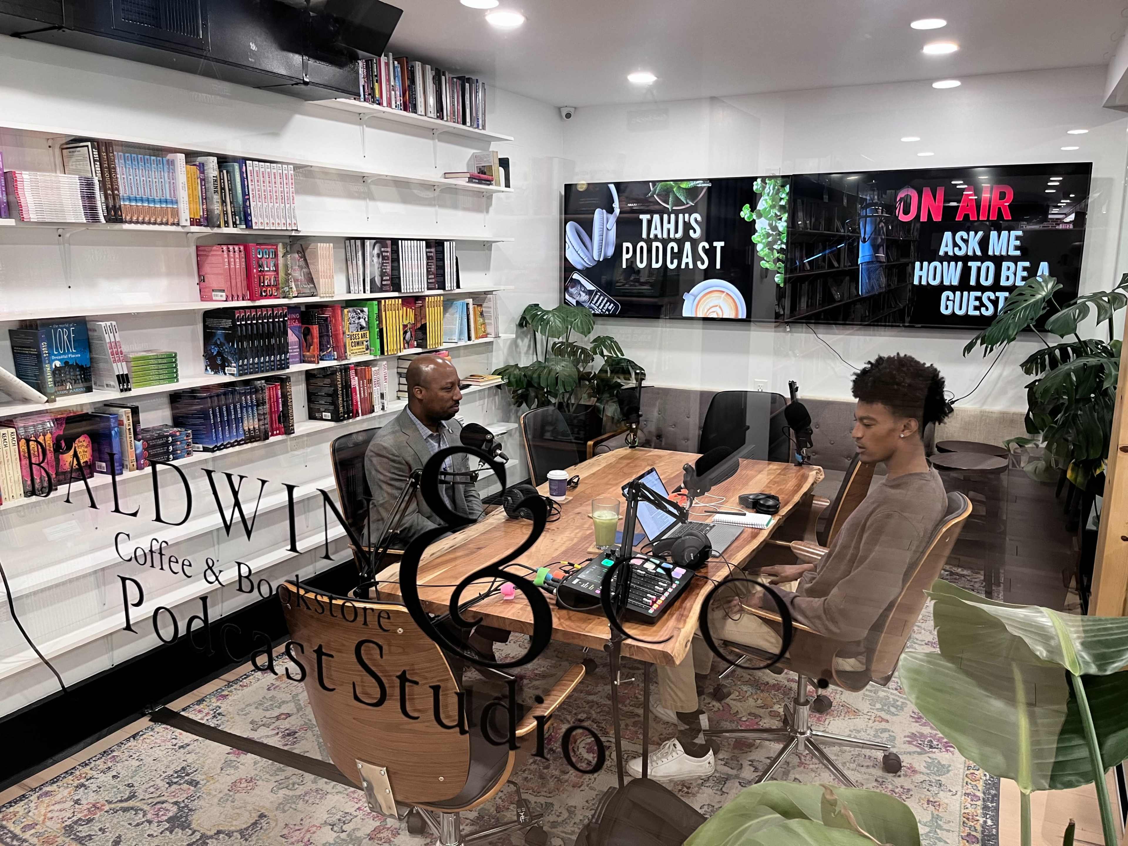 Two individuals sit at a wooden table in a podcast studio, surrounded by bookshelves and large screens displaying the podcast title and "ON AIR" sign.