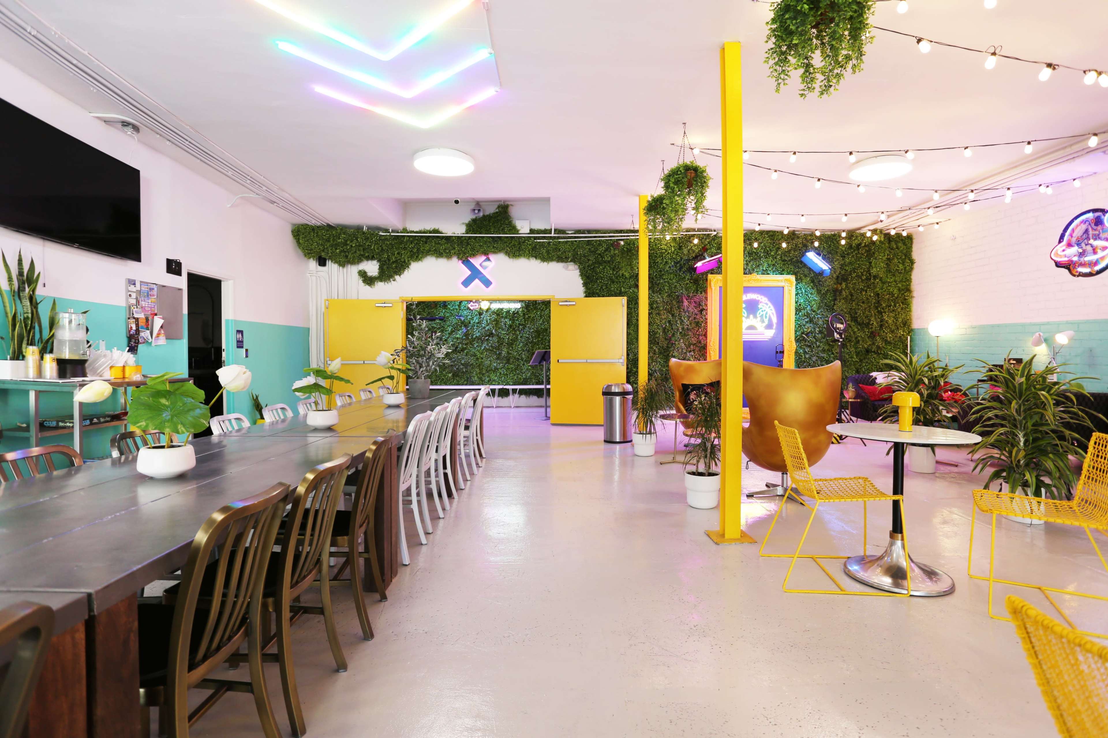 A spacious and colorful coworking space features a long communal table, vibrant decor, and plants along the walls, creating distinct areas for collaboration and relaxation.