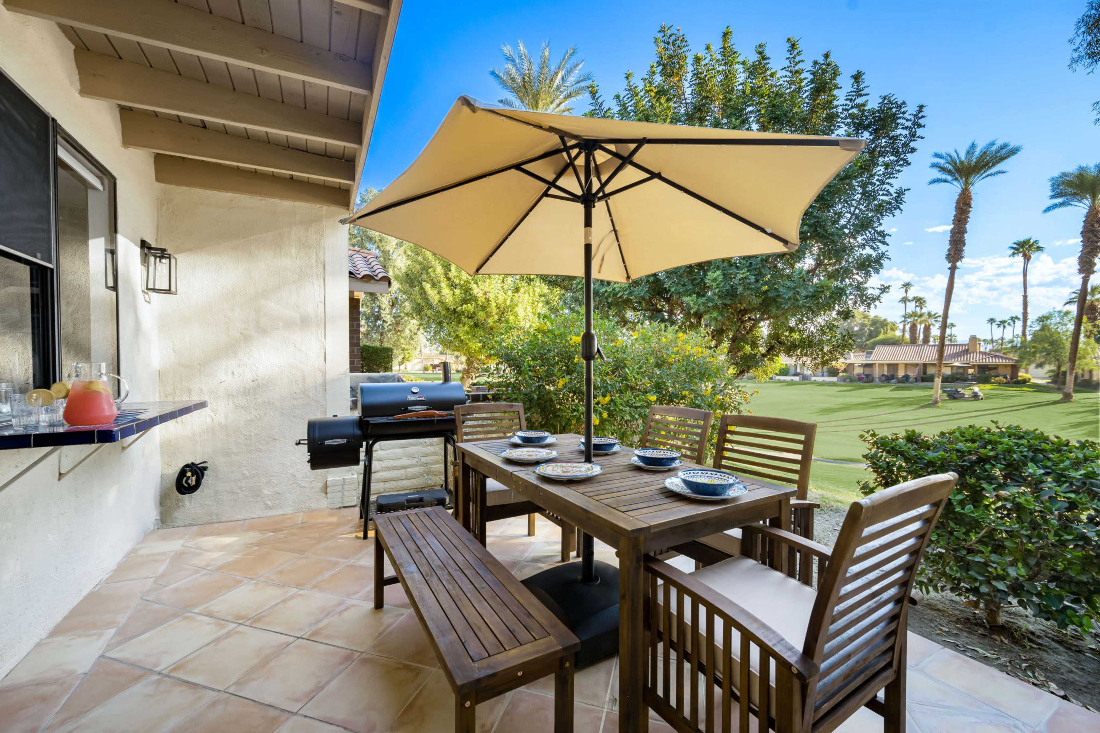 A patio with a wooden dining table and chairs, an umbrella for shade, and a grill, surrounded by a view of palm trees and a green lawn.