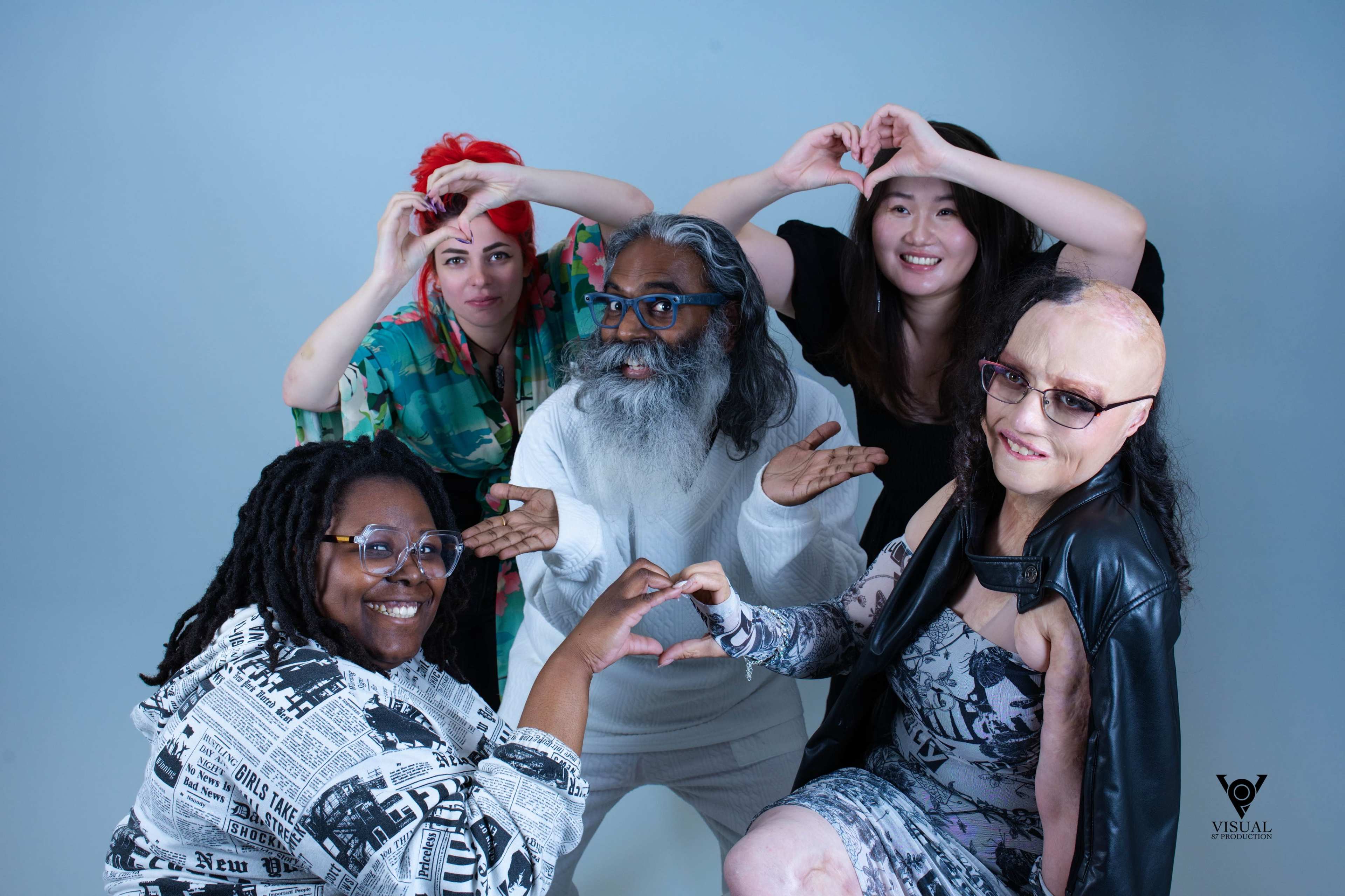 Five individuals are posing together against a light blue background, forming heart shapes with their hands.