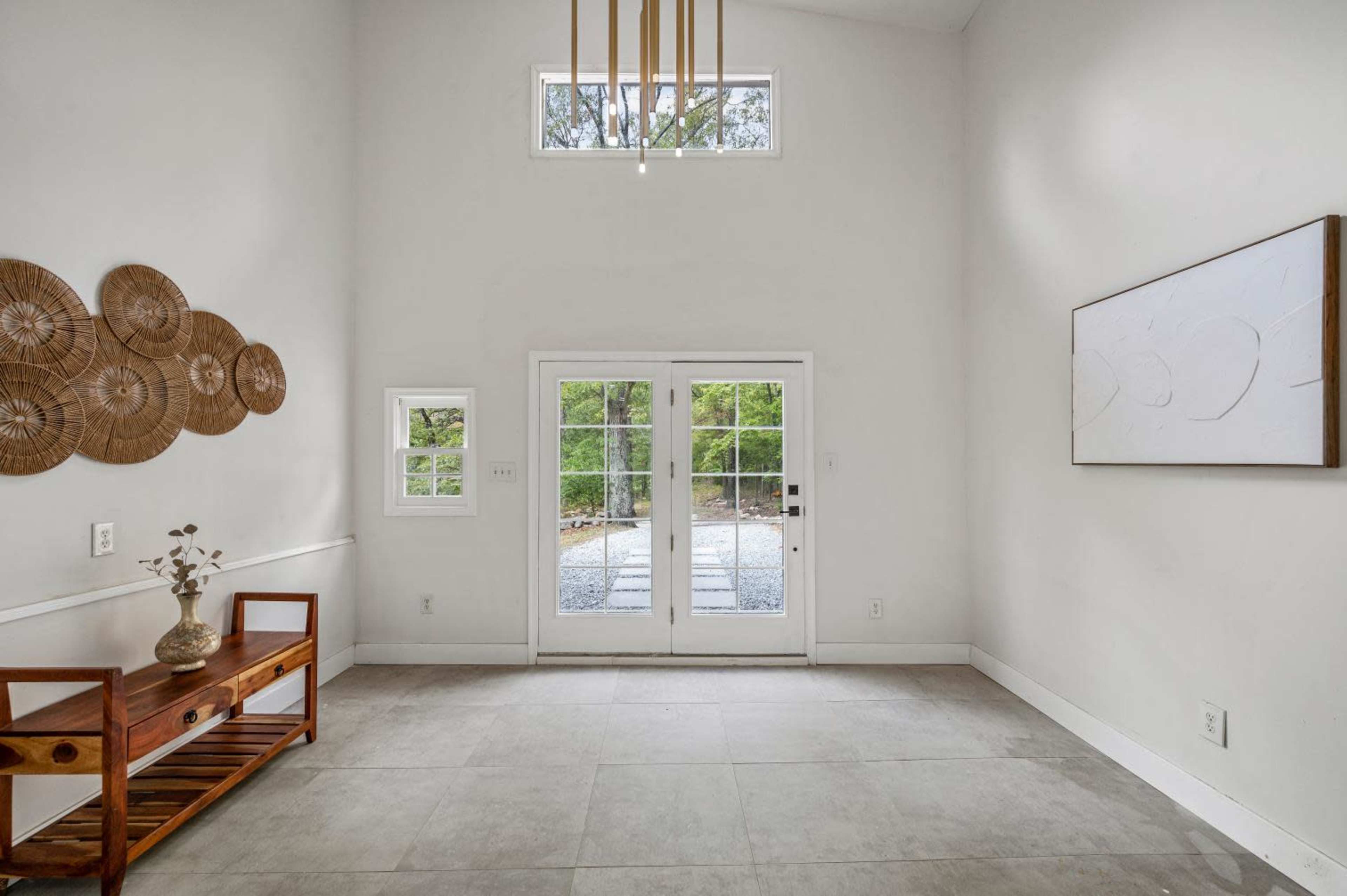 A bright, airy room with a high ceiling features double doors opening to an outdoor space and decorative wall hangings made of natural fibers.