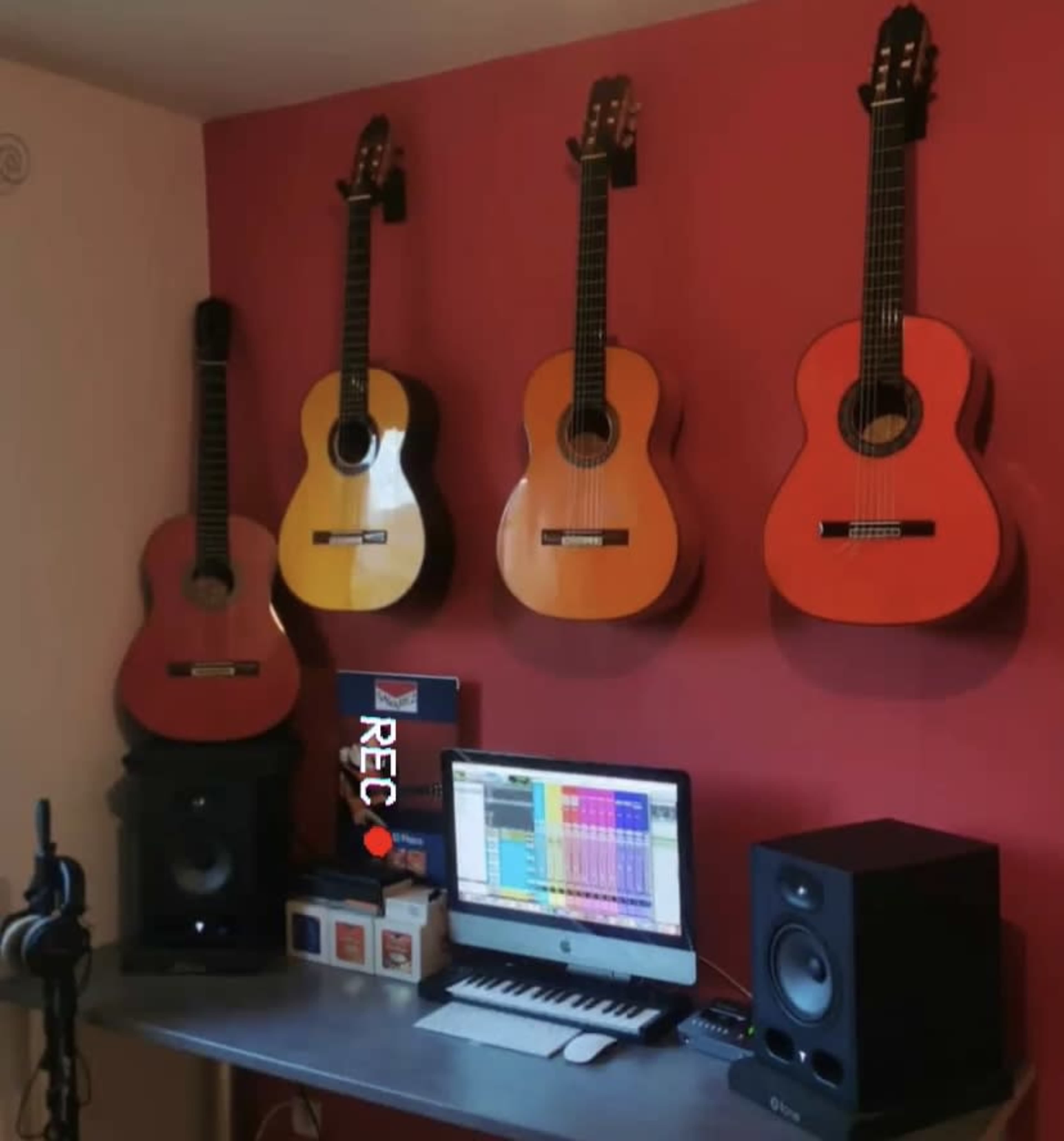 The image shows a home recording studio setup featuring four guitars mounted on a red wall, with a computer and audio equipment on a desk below.