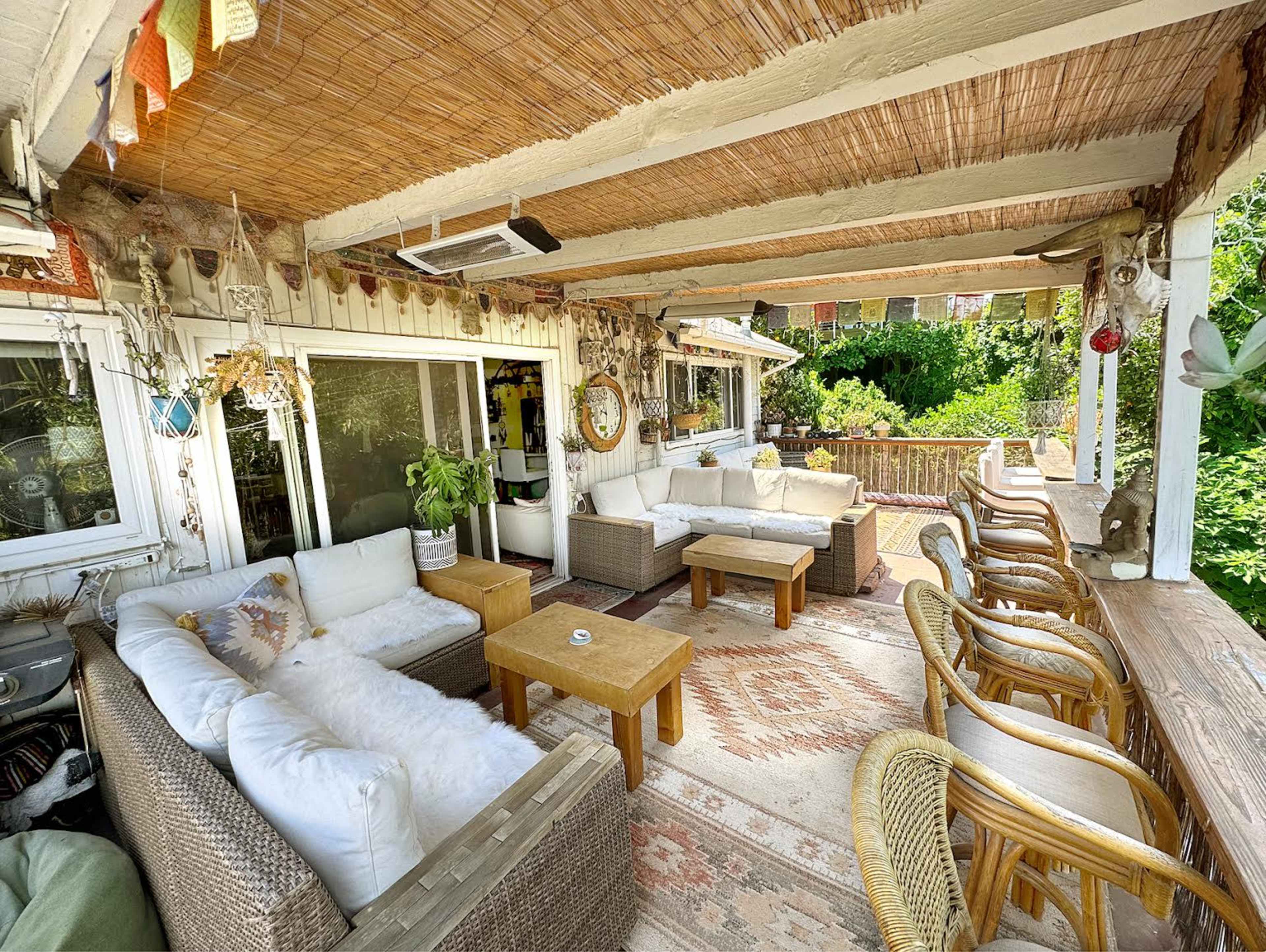 A cozy outdoor seating area with wicker furniture, a wooden table, and decorative elements surrounded by greenery.