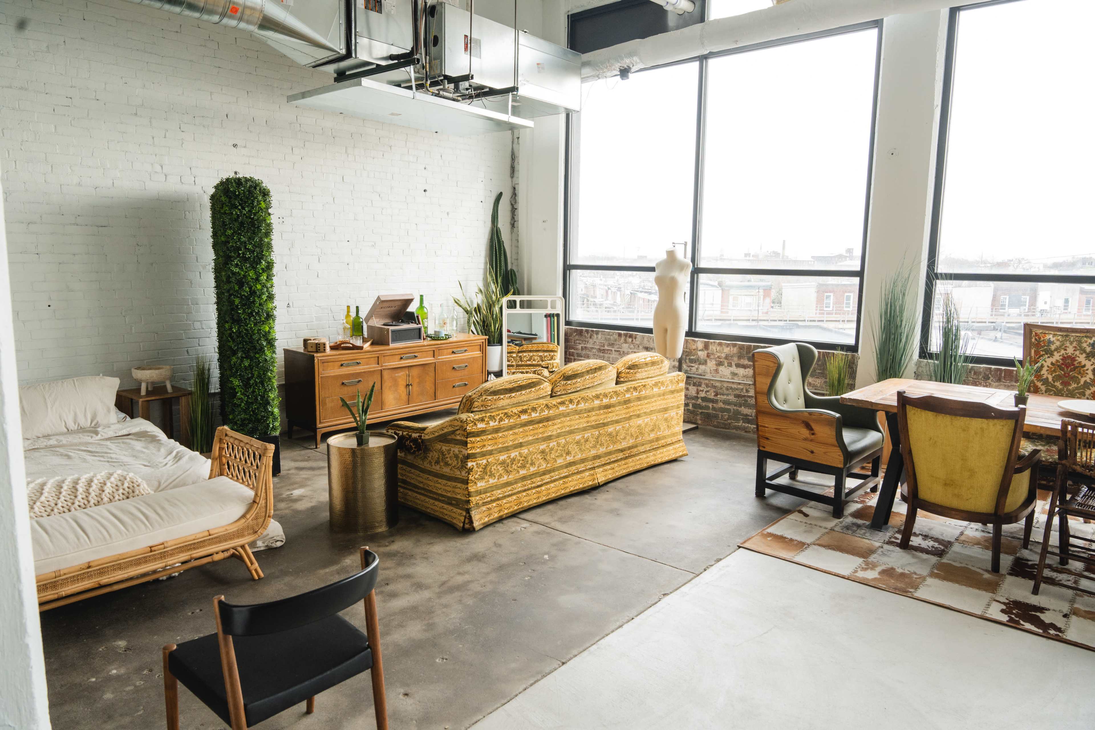 A spacious room features a large window with urban views, a bed, a wooden storage unit, and various seating arrangements, including a mannequin and indoor plants.