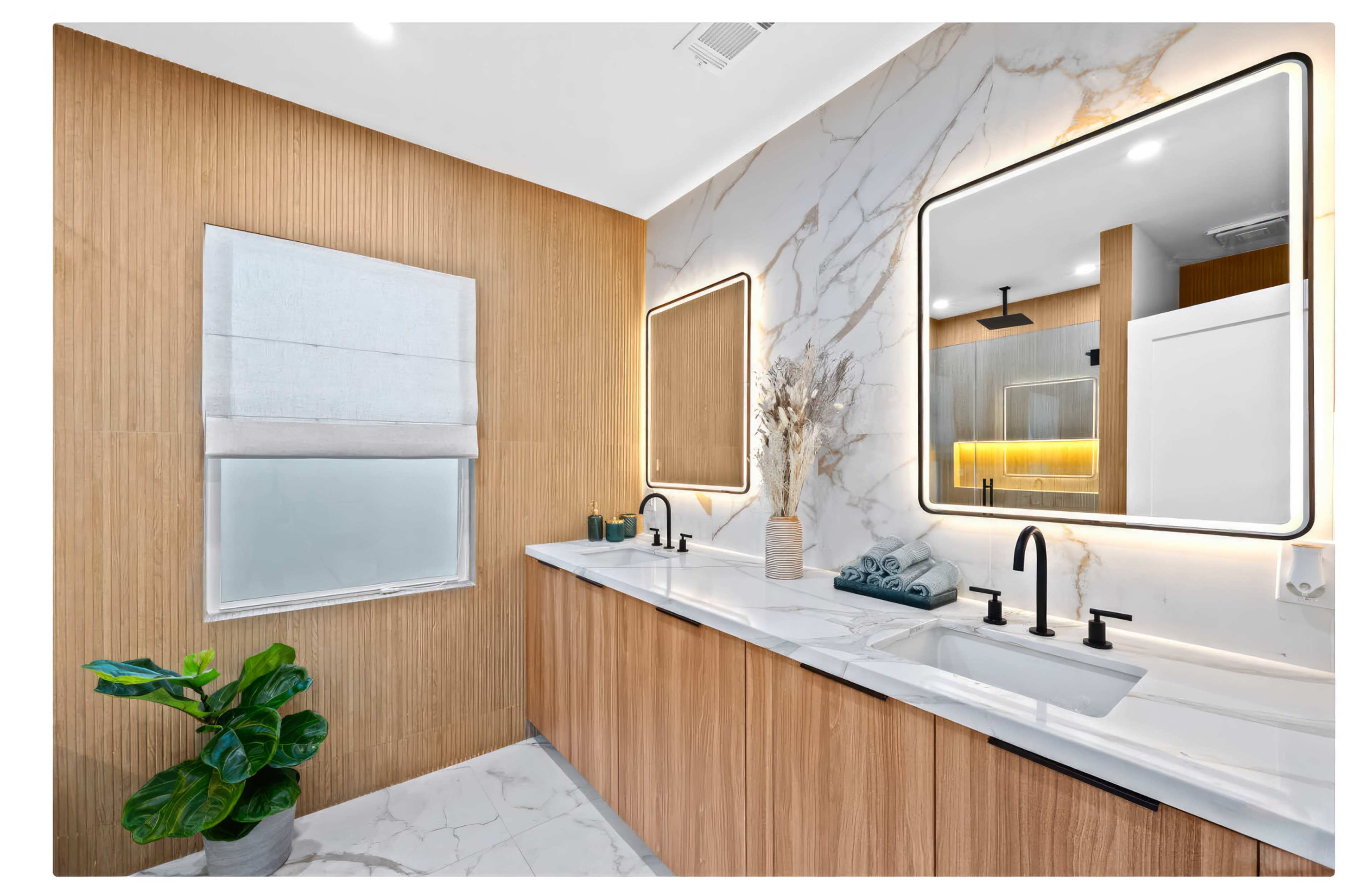 The bathroom features a double vanity with modern rectangular mirrors, light wood accents, and a large marble wall, complemented by a window dressed with a fabric shade and potted plant.
