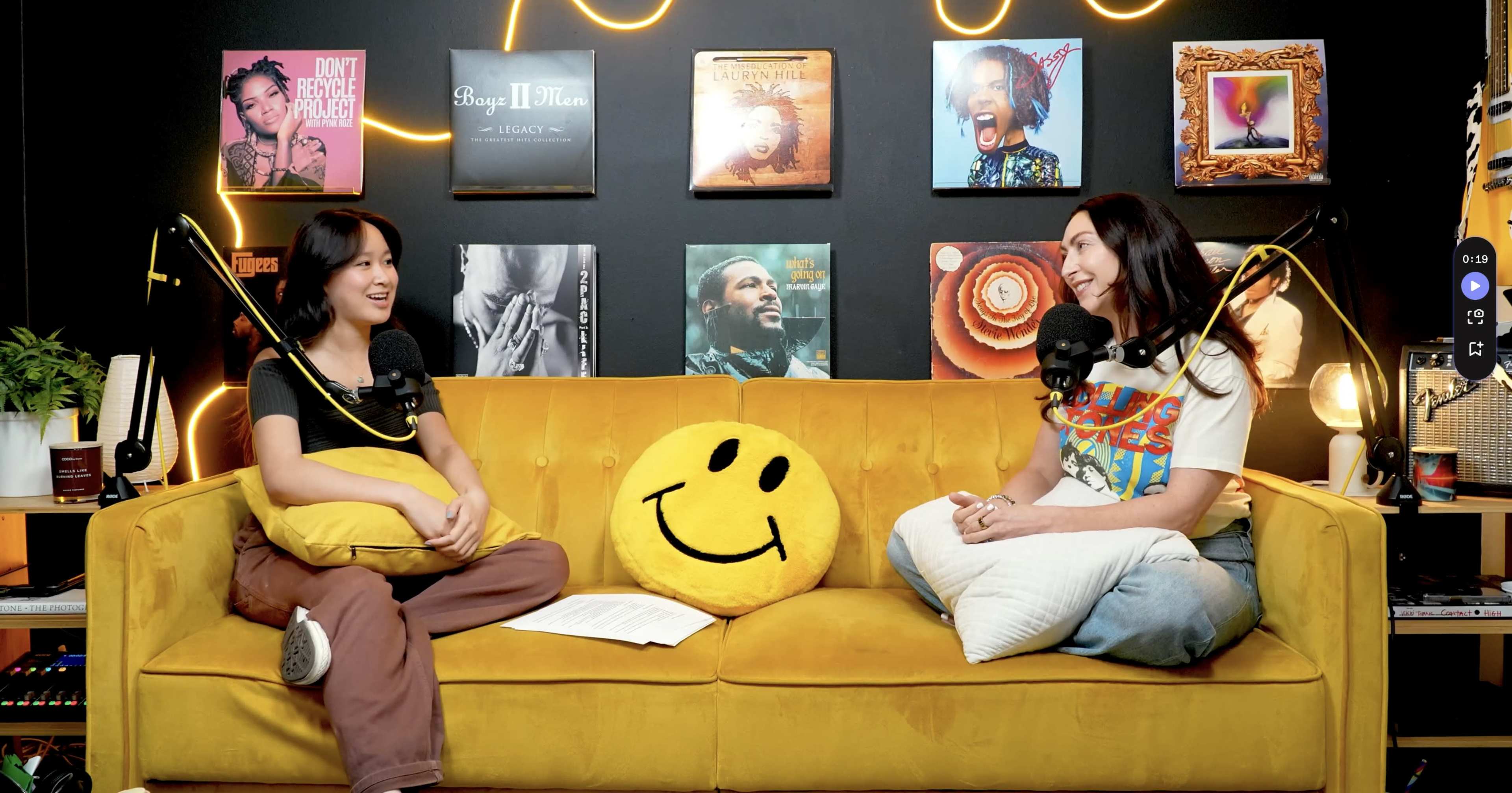 Two women sit on a yellow couch adorned with a large smiley face pillow, engaged in conversation in front of a wall decorated with framed album covers.