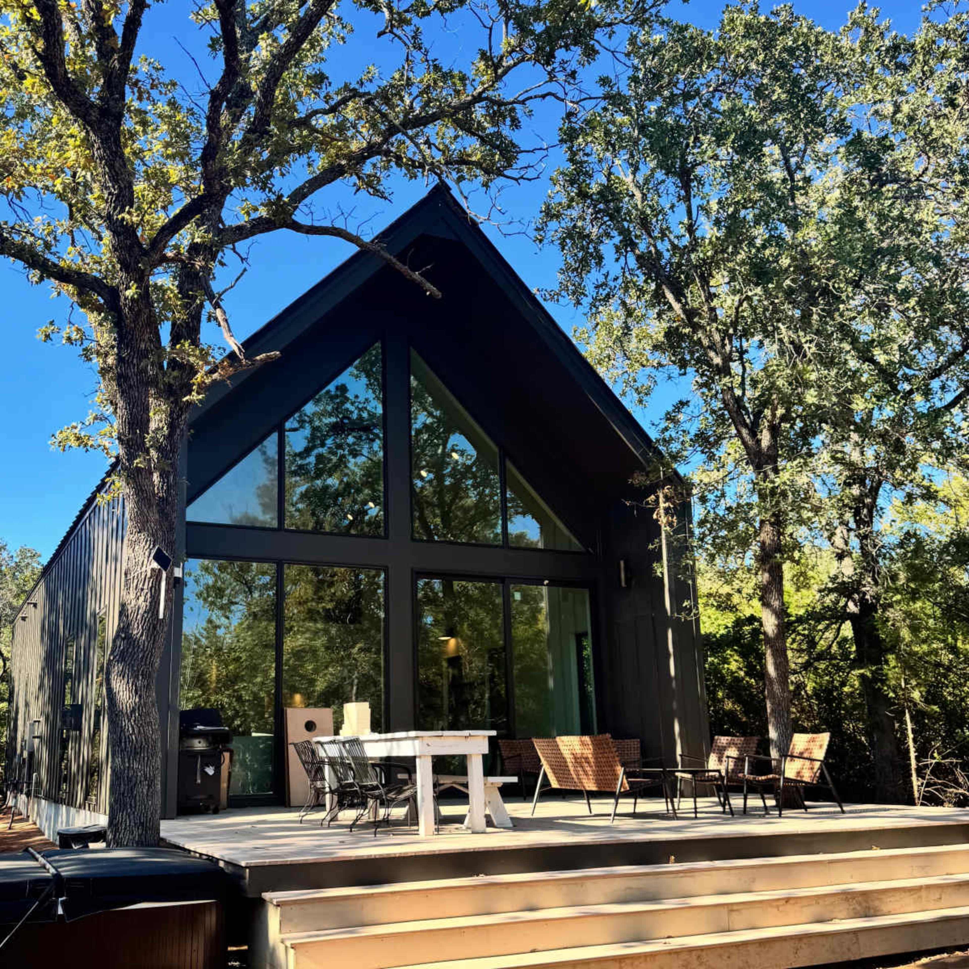 The image shows a modern, triangular-roofed cabin surrounded by trees, featuring large glass windows and a wooden deck with outdoor furniture.