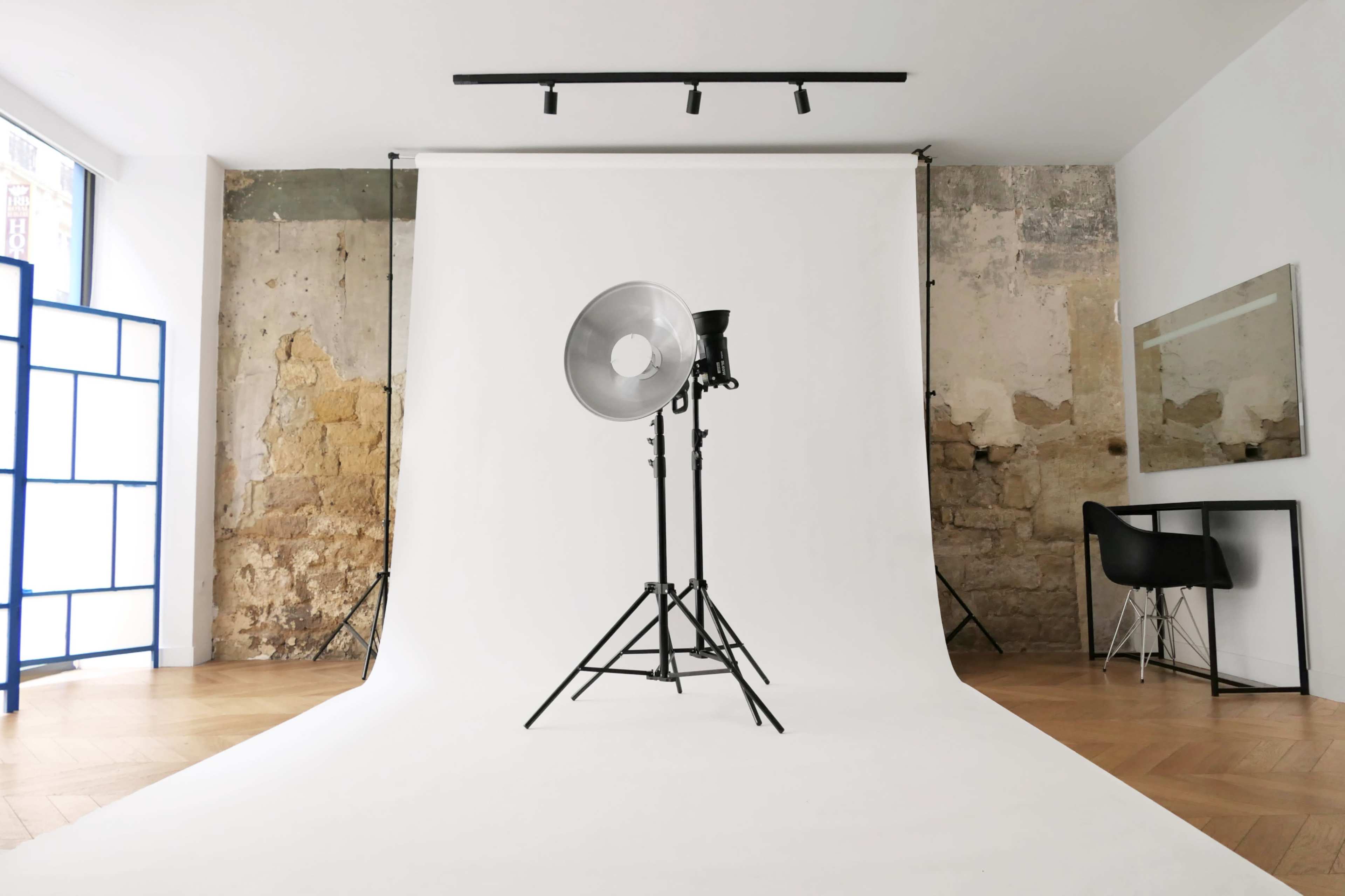 A photography studio setup with a white backdrop, a large light fixture on a stand, and a chair beside a mirror against a textured wall.