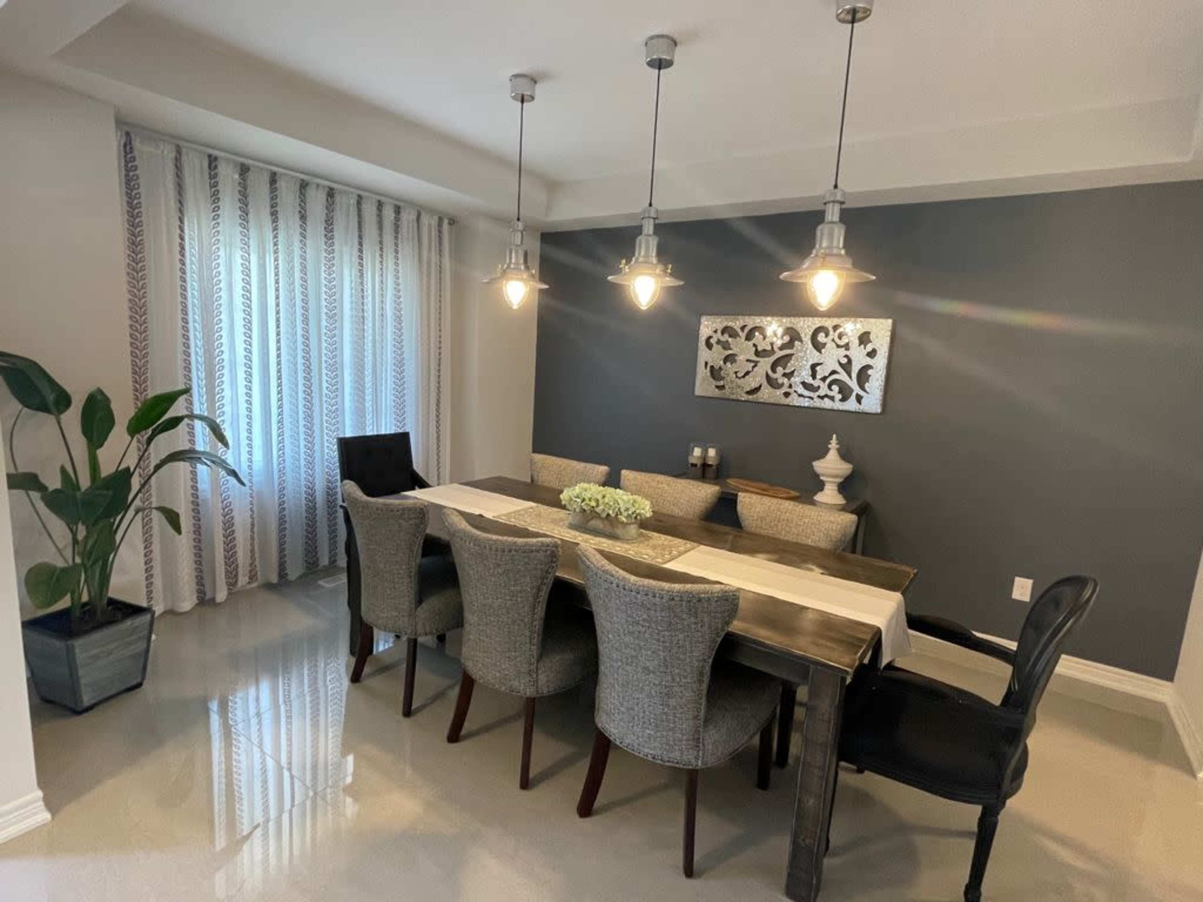 A dining area features a long table surrounded by gray and black chairs, with pendant lights hanging above and a plant in the corner.