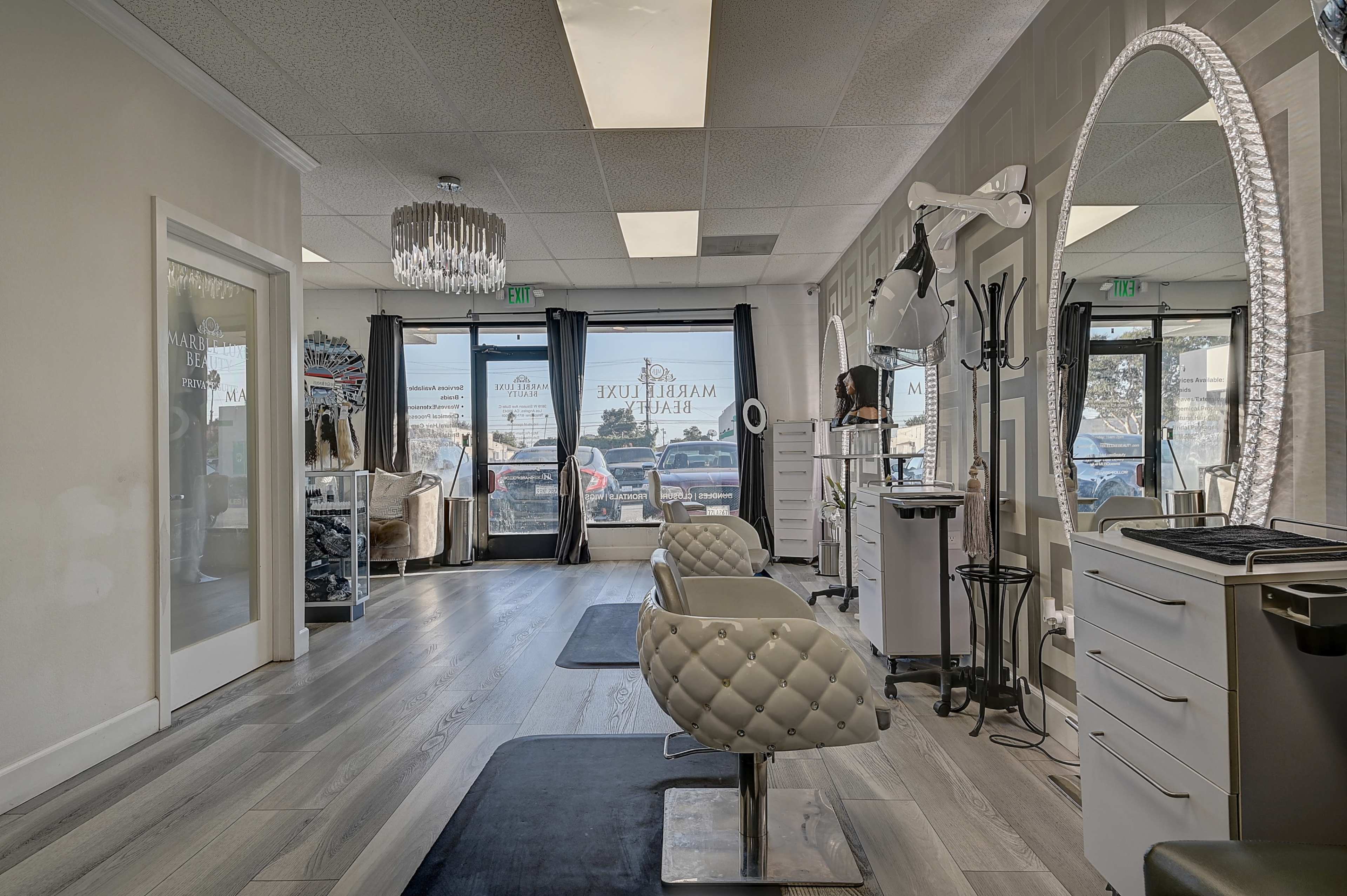 The interior of a boutique salon featuring two white stylized chairs, a large round mirror, and a modern decor with clothing displays visible through the front windows.