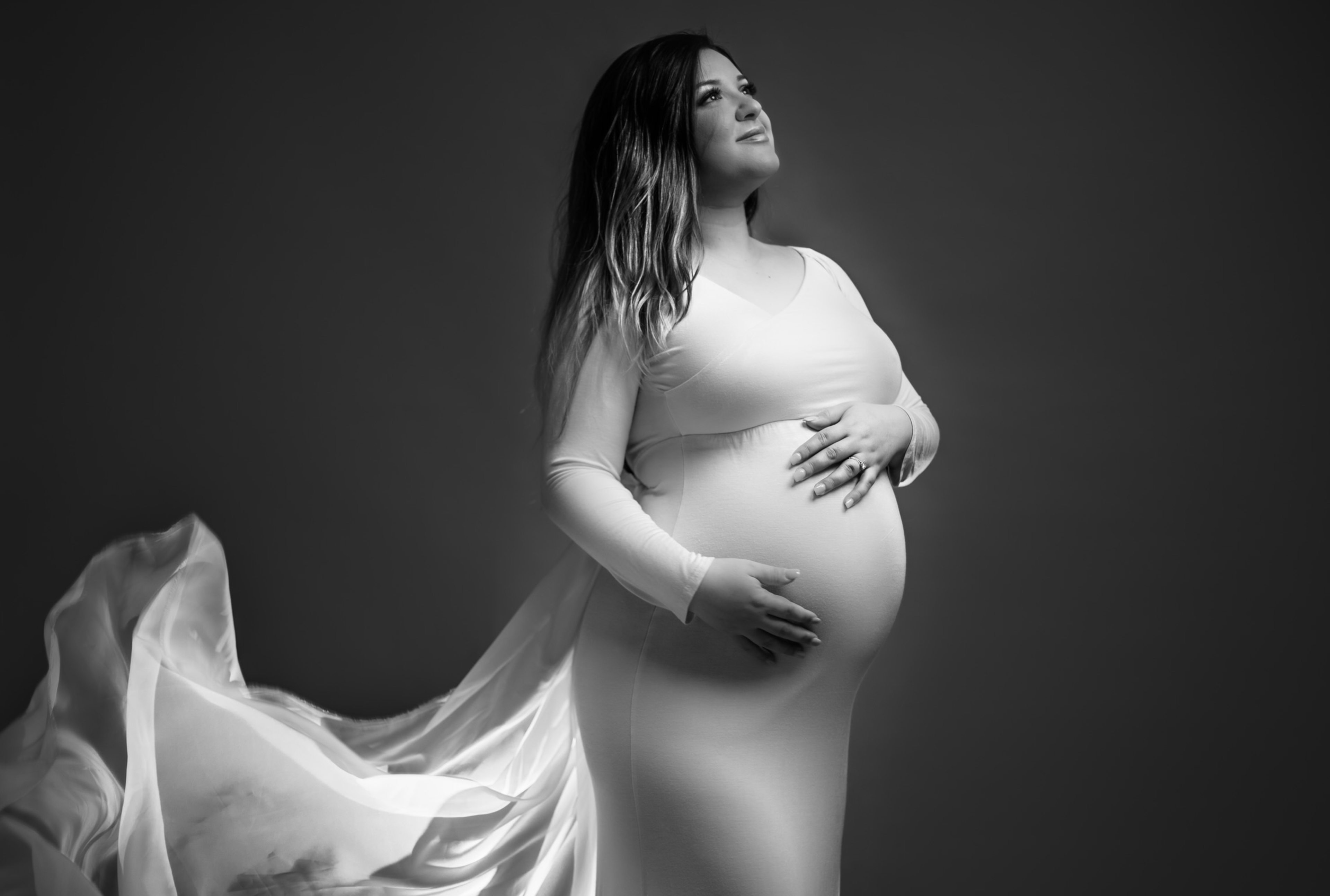 A maternity photoshoot captures a pregnant woman in a white gown in black and white.